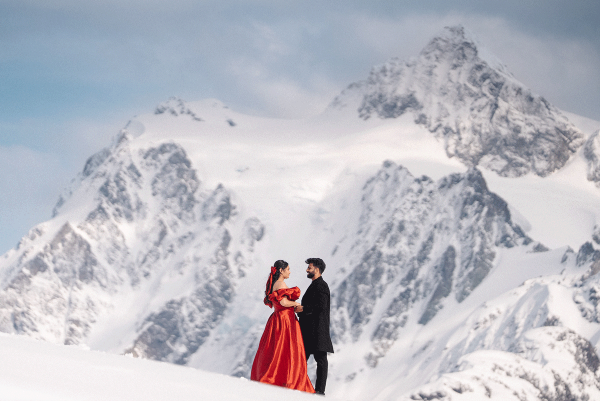 Couple in formal wear stands atop a snowy mountain peak with dramatic snow-capped mountains in the background.