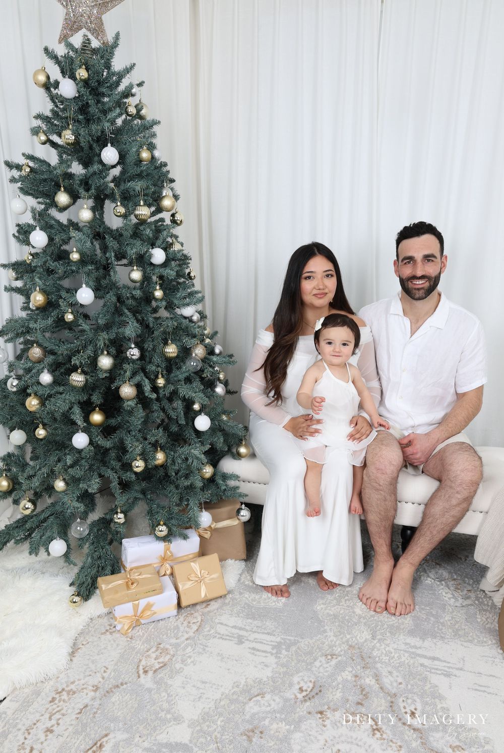 A family poses together in white outfits next to a decorated Christmas tree with white ornaments and wrapped presents below.
