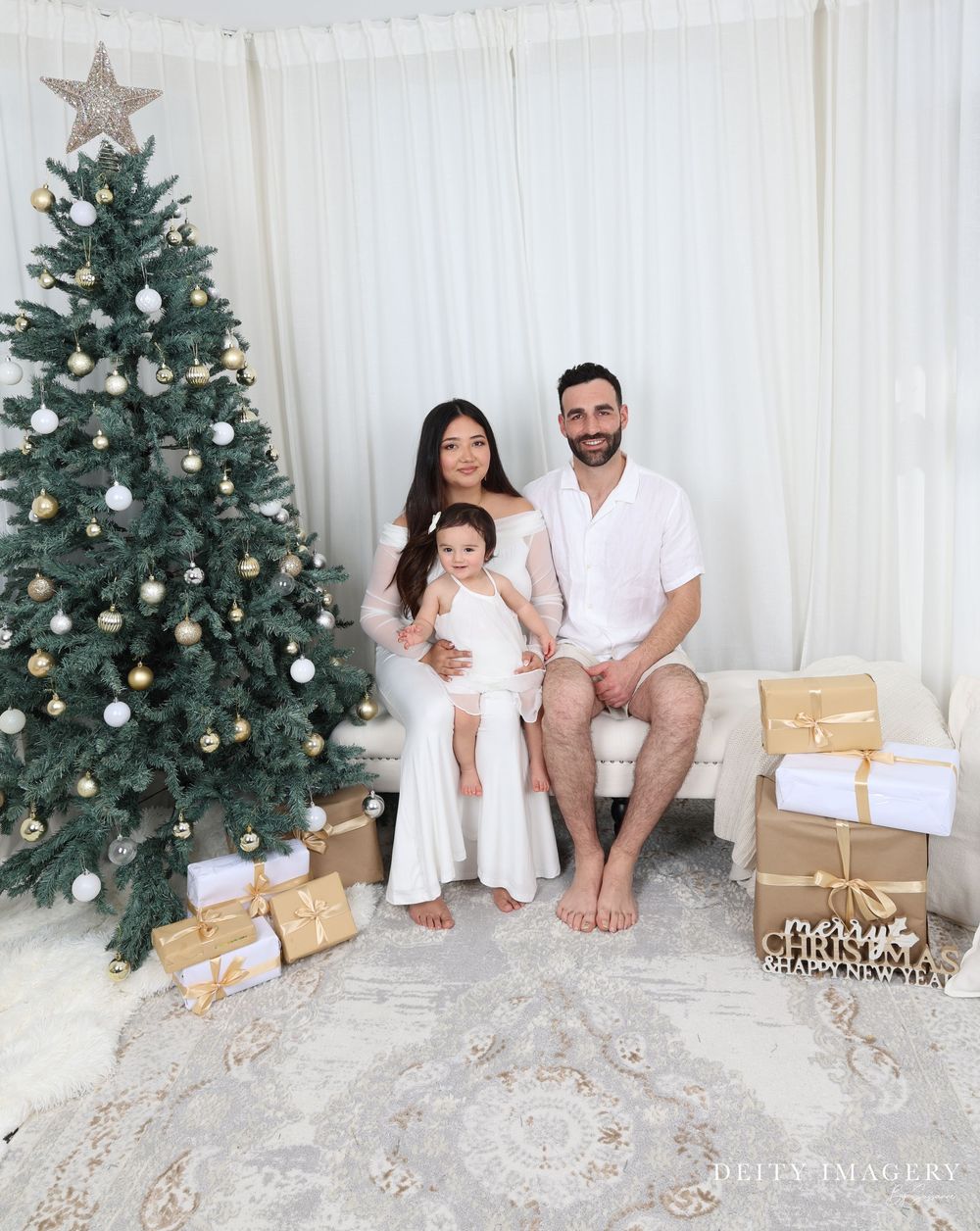 Family sitting near decorated Christmas tree with wrapped presents in white toned photoshoot sequence.