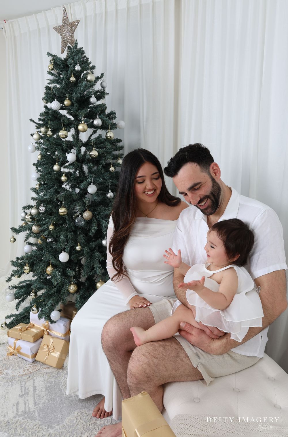 A family shares tender holiday moments in white outfits by a decorated Christmas tree with gold wrapped presents.