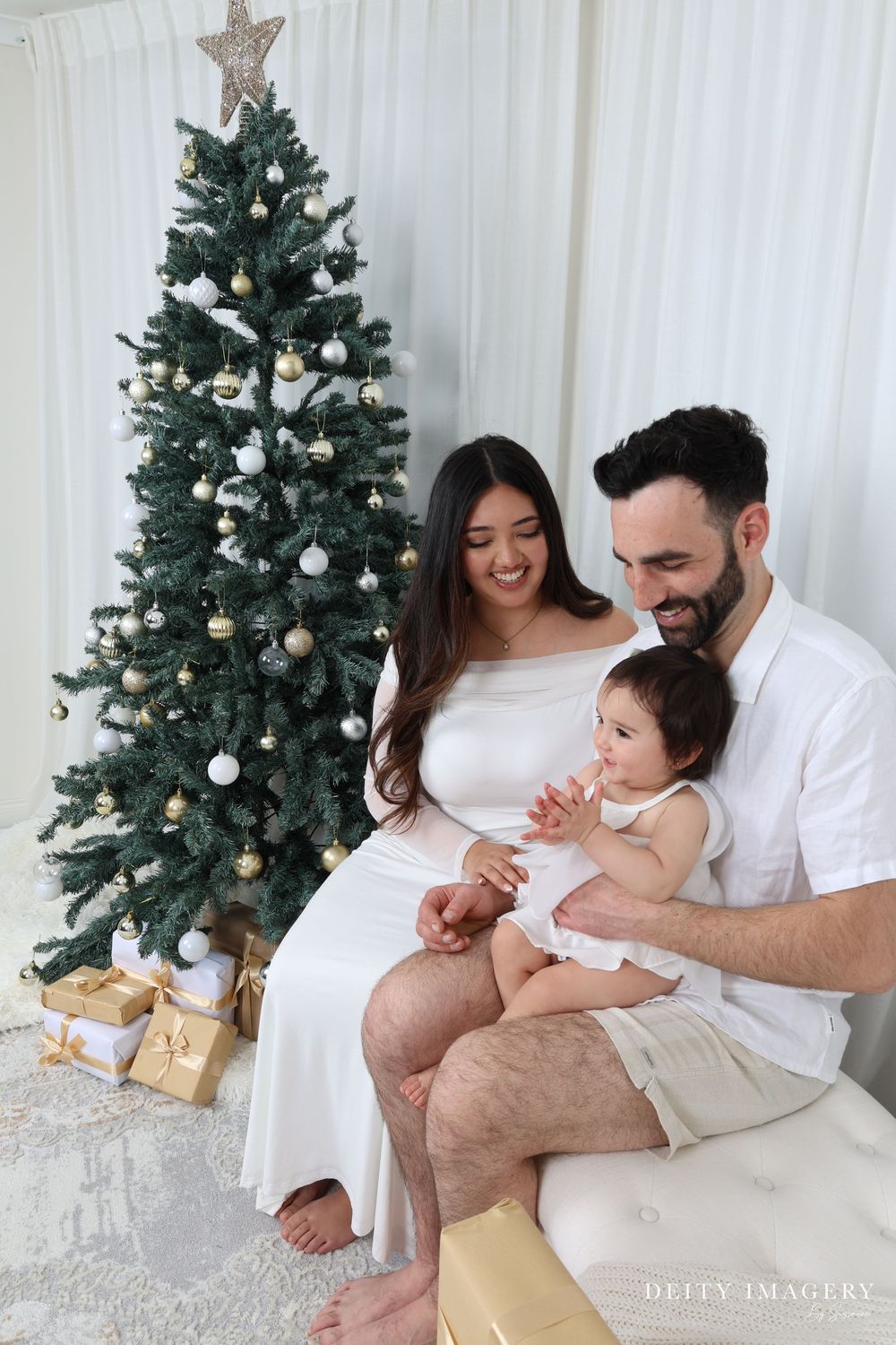 A family poses for intimate Christmas portraits next to a decorated tree with gold presents beneath it.