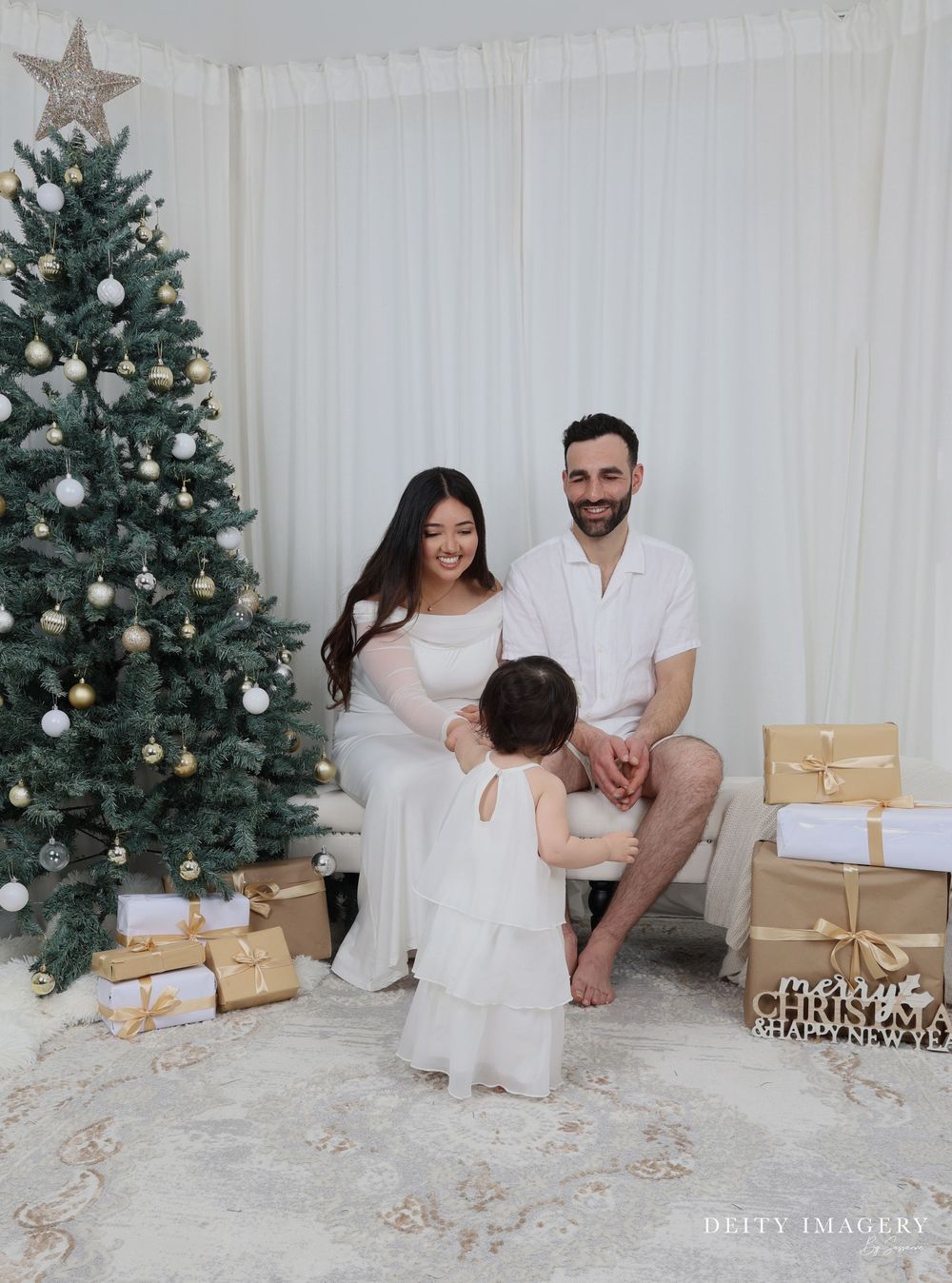 A decorated Christmas tree stands next to wrapped presents as a family in white outfits poses for a holiday photo.