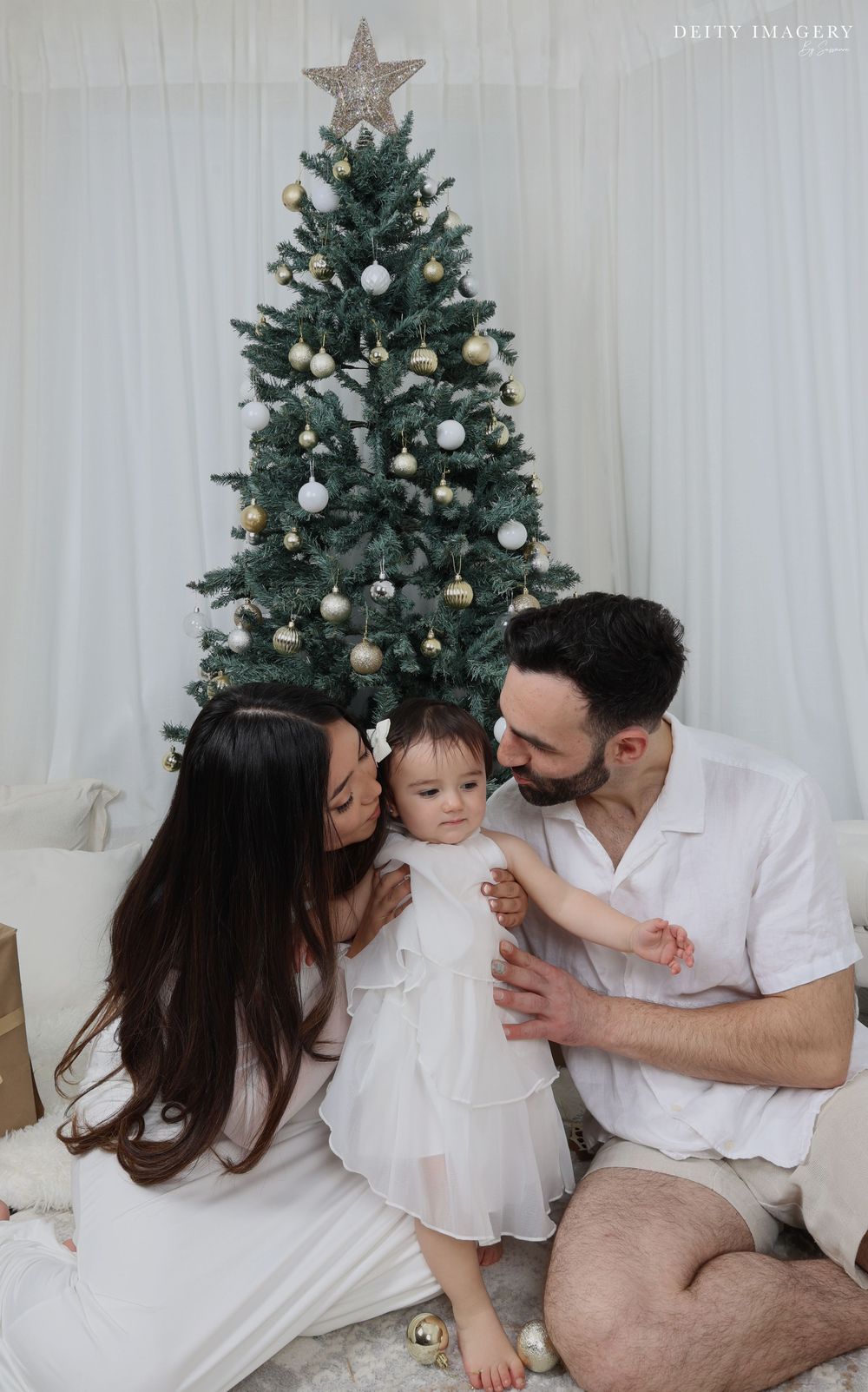 Family sharing tender holiday moments by decorated Christmas tree in white matching outfits in cozy home setting.