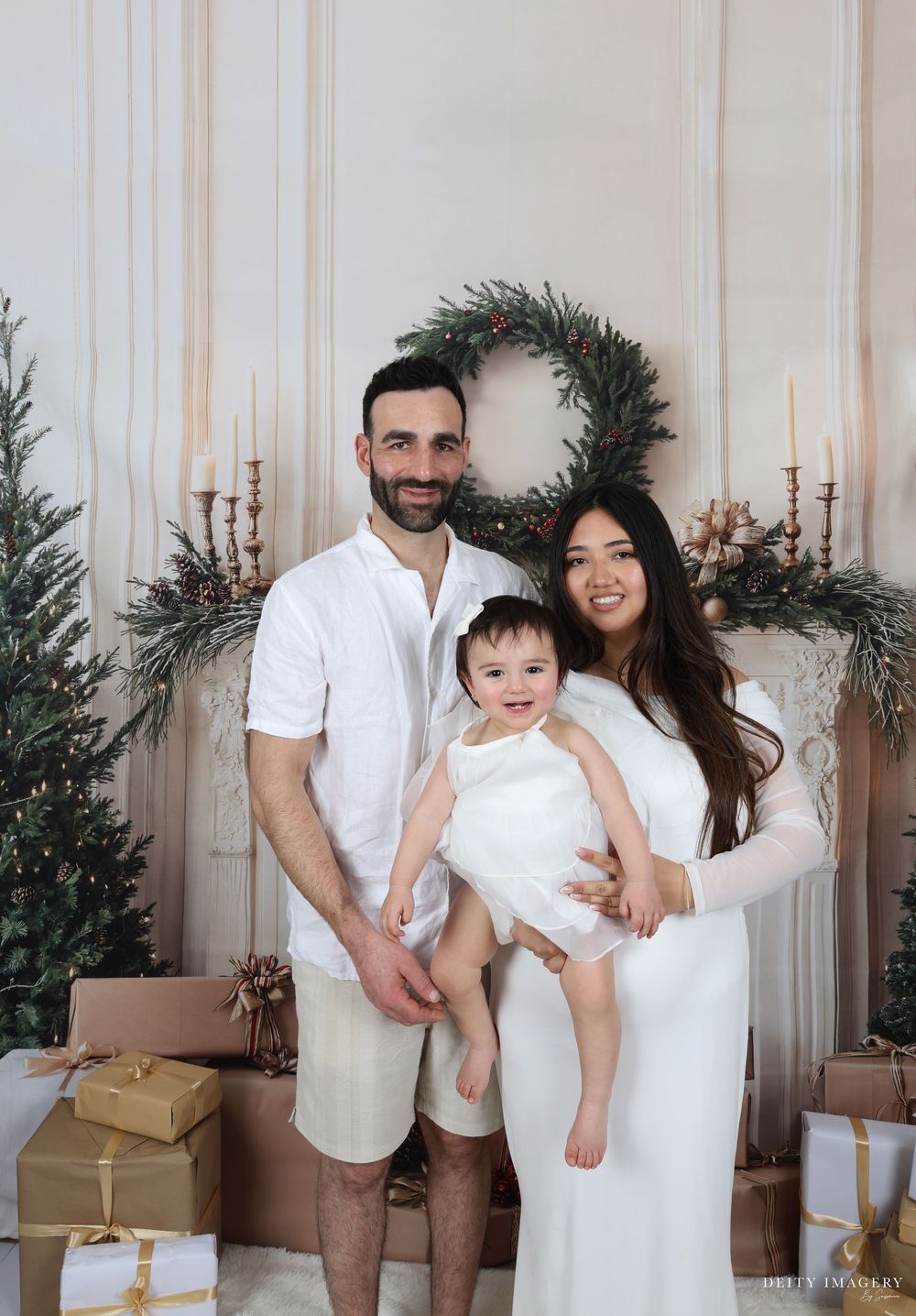 A family poses together in front of a holiday wreath and decorative backdrop with wrapped presents at their feet.