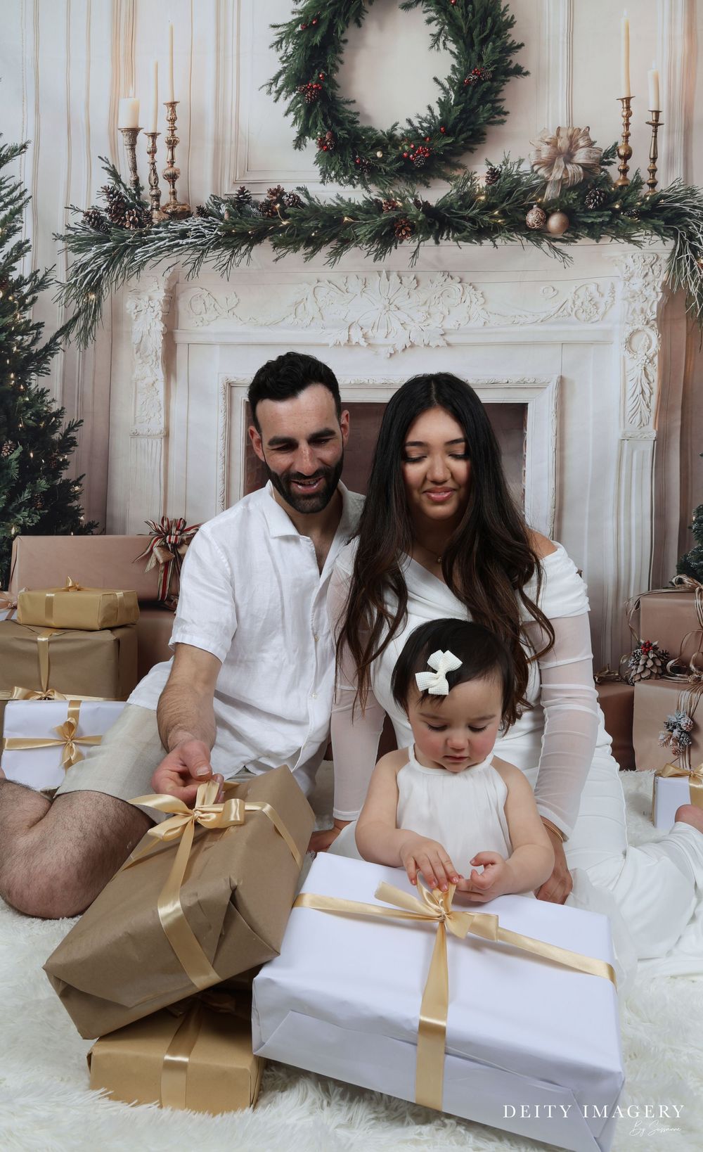 Holiday portrait session shows couple in white outfits opening gifts on floor with Christmas decor and wreaths above.