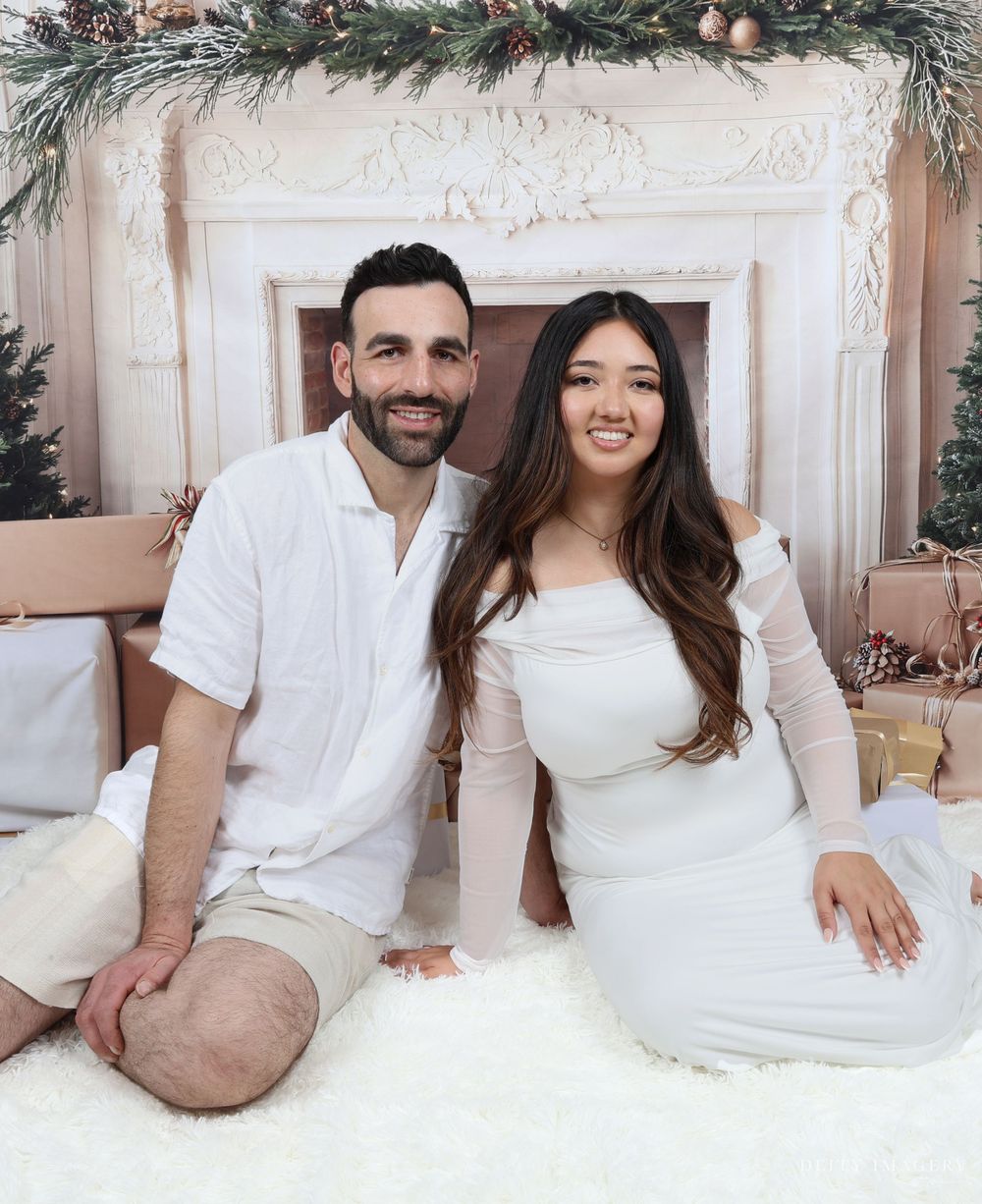 A cozy holiday photo featuring two people in white outfits sitting by a decorated fireplace with garland and festive decor.