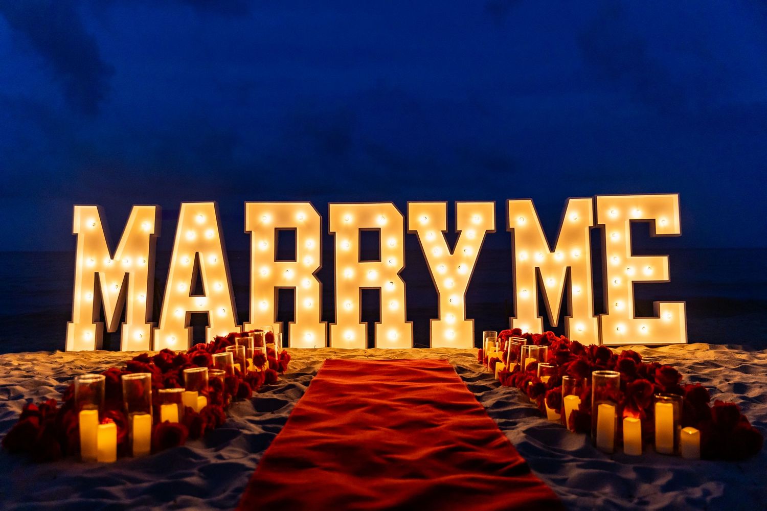 Romantic candle-lit marriage proposal setup with glowing letters spelling 'Marry Me' along a beach at sunset.