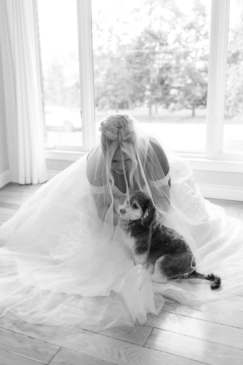 A person in a flowing white wedding dress poses with their black and white dog on a bright sunlit floor.