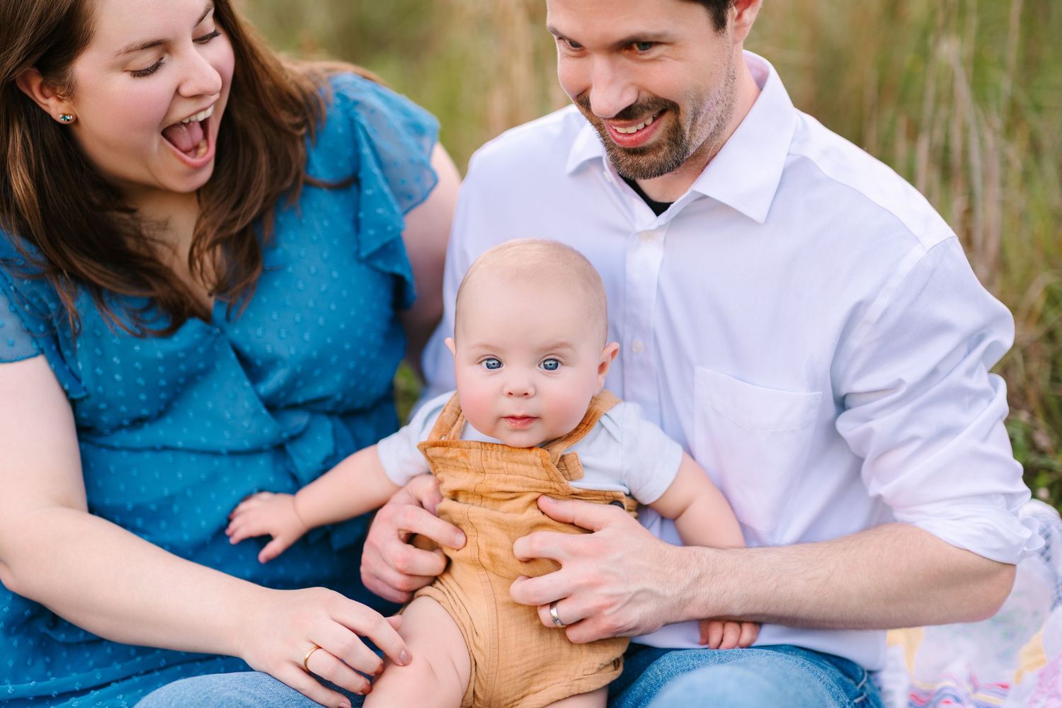 A family in coordinating blue and white outfits laughs together while sitting on a blanket during an outdoor photo session.