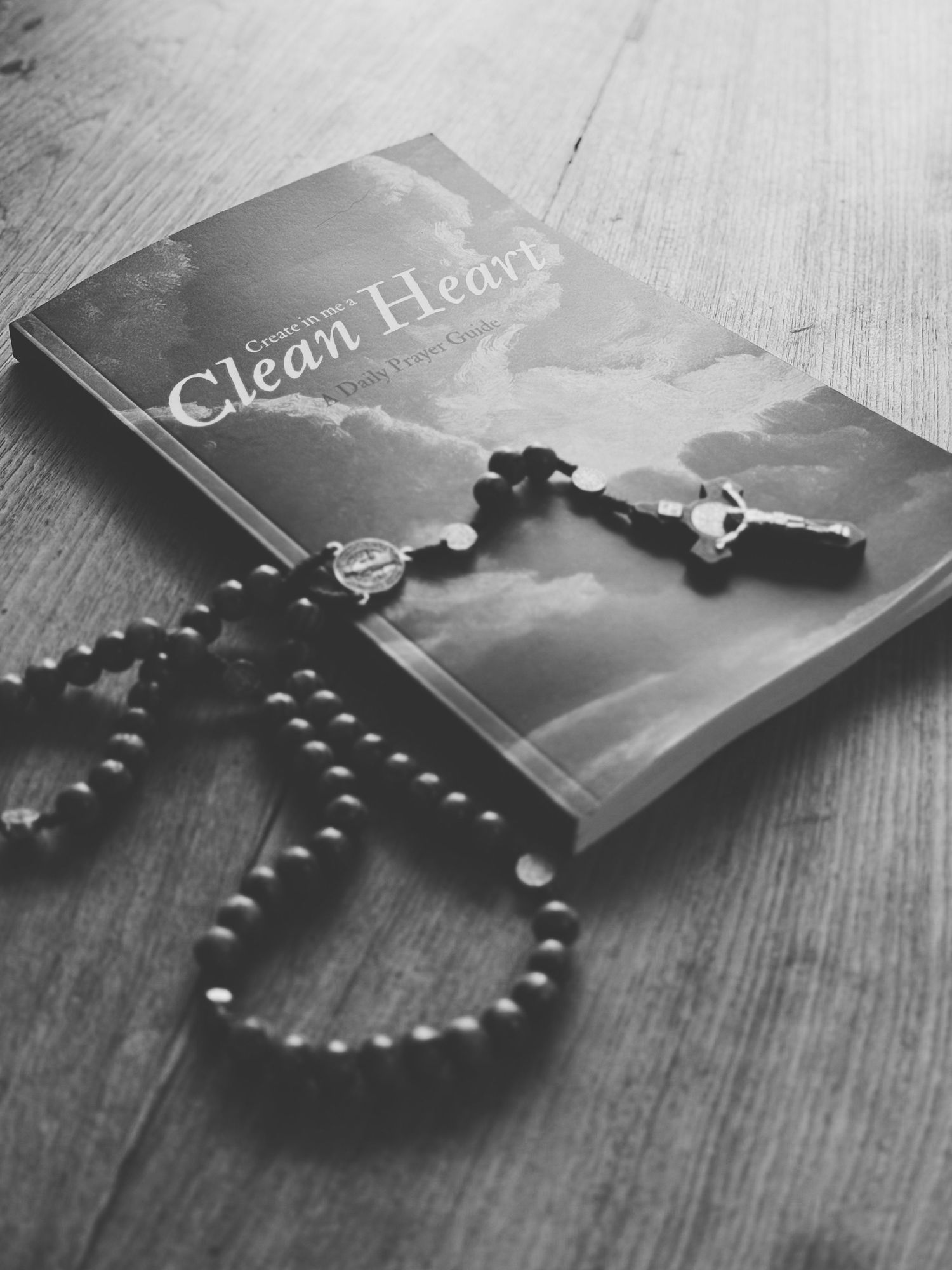 Black and white photo of a rosary necklace arranged in a heart shape with a book on wooden surface.
