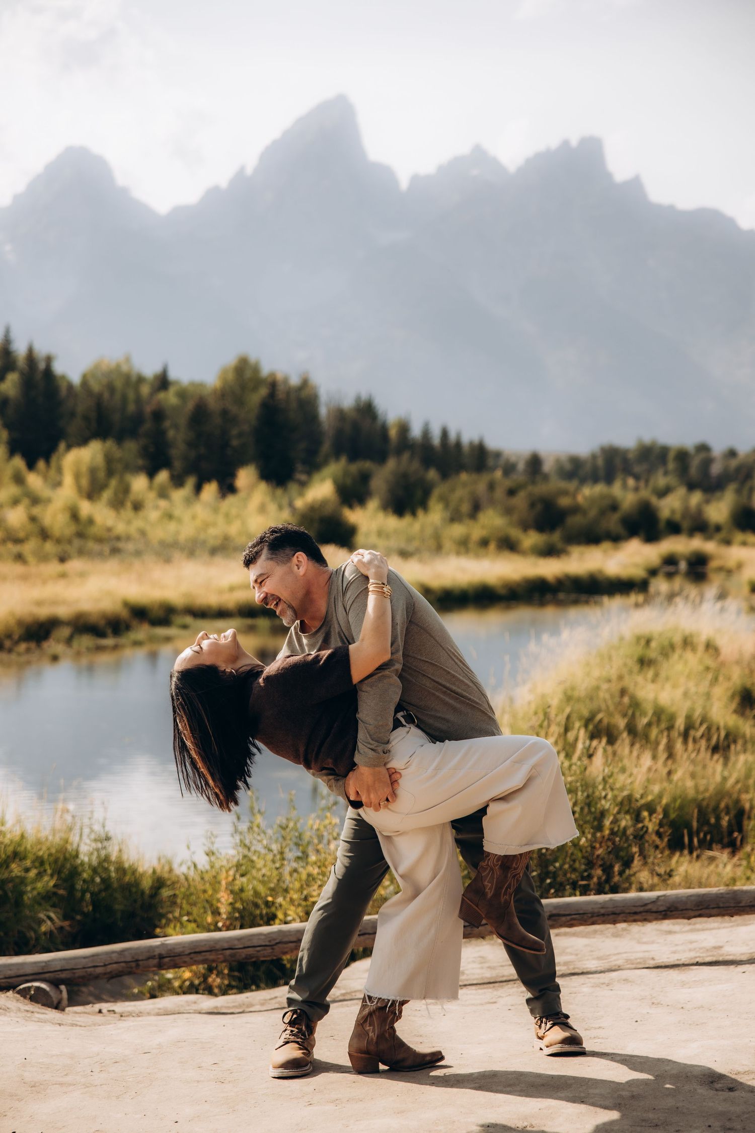 A romantic couple poses playfully near a scenic mountain lake with dramatic peaks in the background.