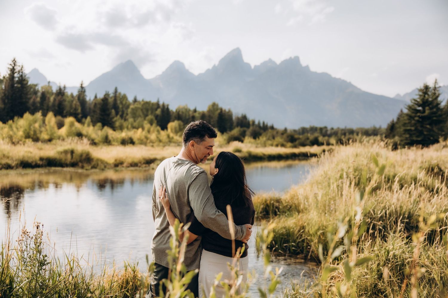 A couple embraces by a serene lake with majestic mountain peaks rising in the background during golden hour.