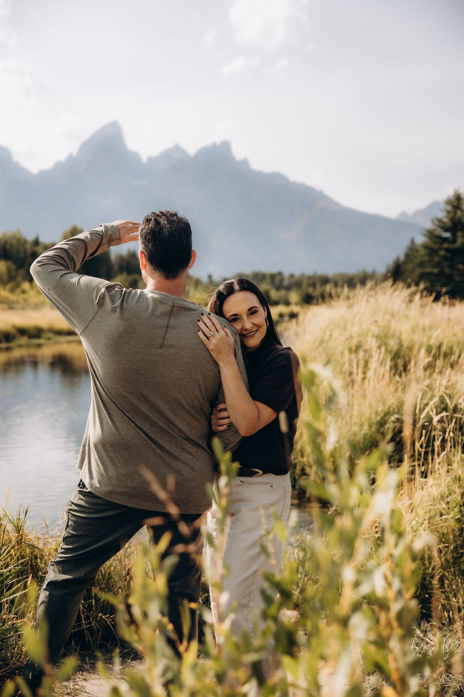 A couple shares a romantic moment in a grassy meadow with mountain peaks and a lake in the background.