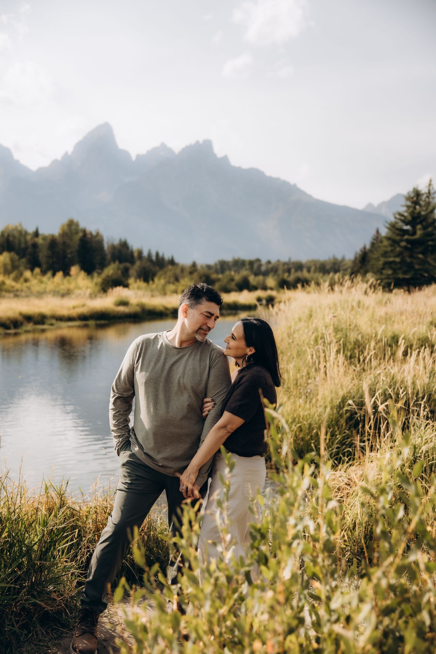 A couple shares tender moments in a scenic mountain landscape with tall grasses and a lake at Grand Teton National Park.