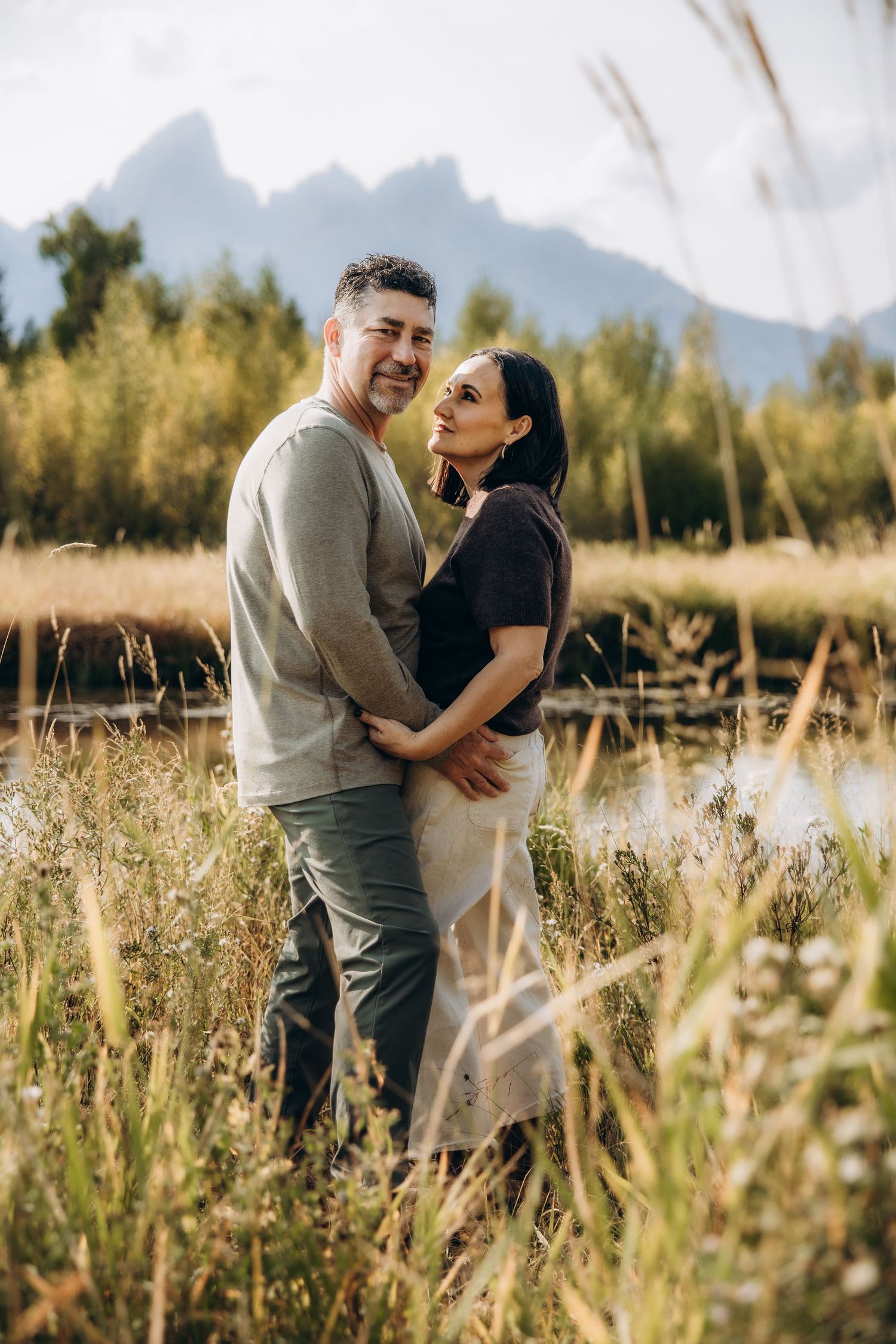 A couple shares tender moments together in a sunny mountain meadow with tall grass and mountain peaks in the background.