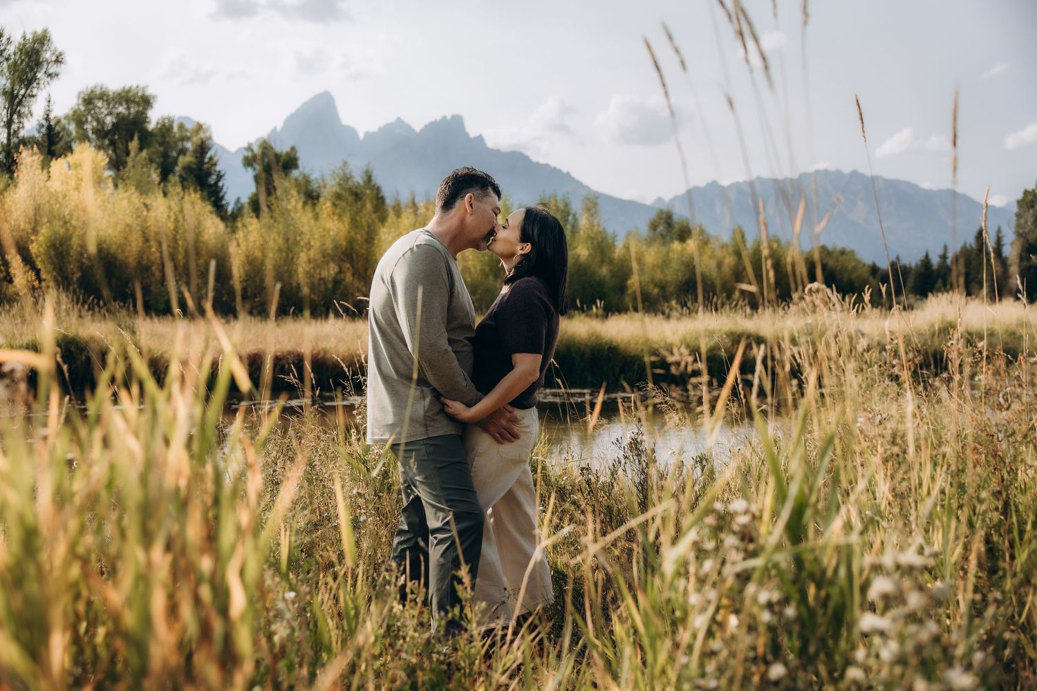 A couple embraces in tall grass with majestic mountains in the background at golden hour.
