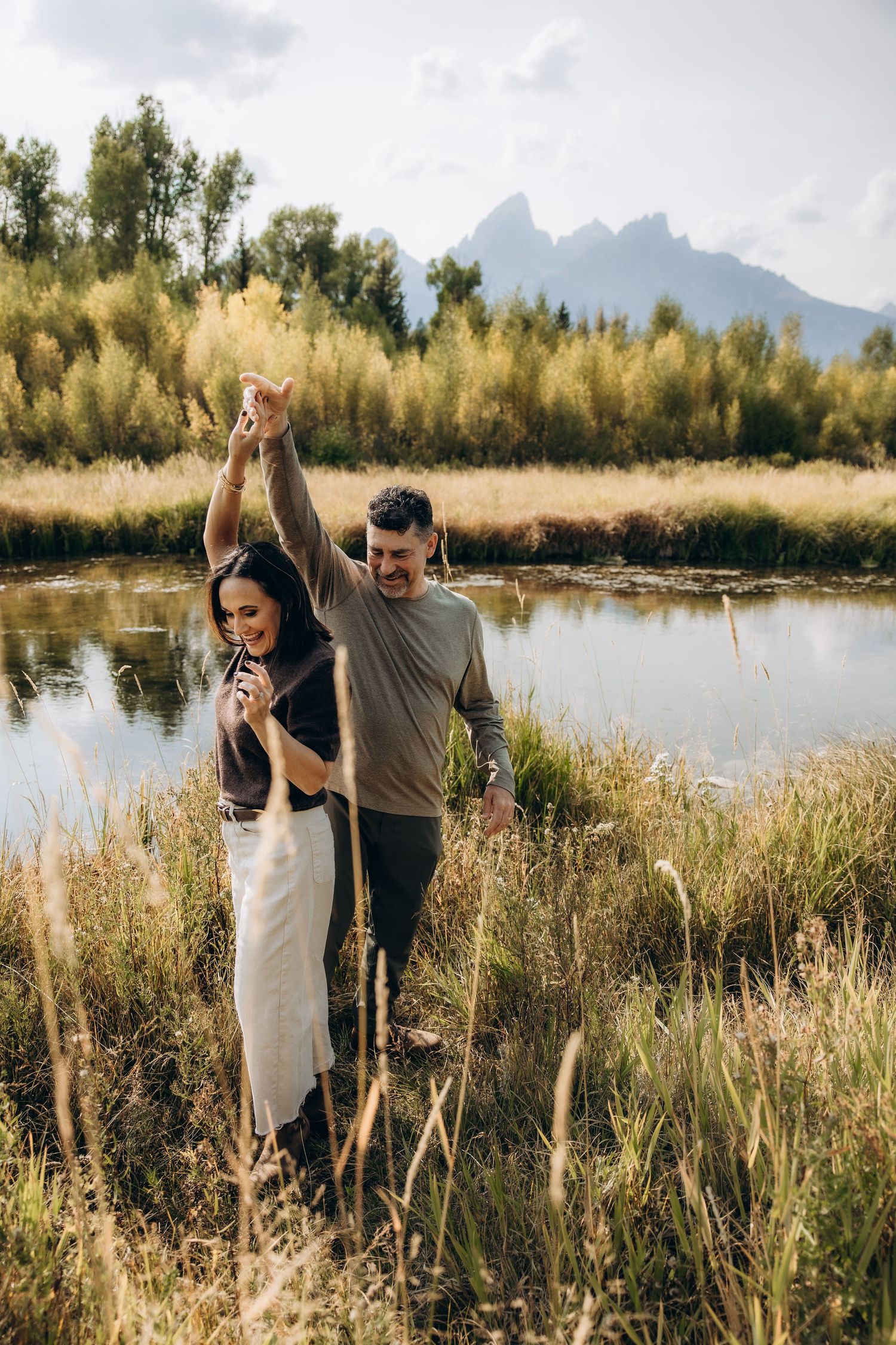 A couple embraces in a scenic outdoor setting with tall grass and a tranquil pond against a mountain backdrop.