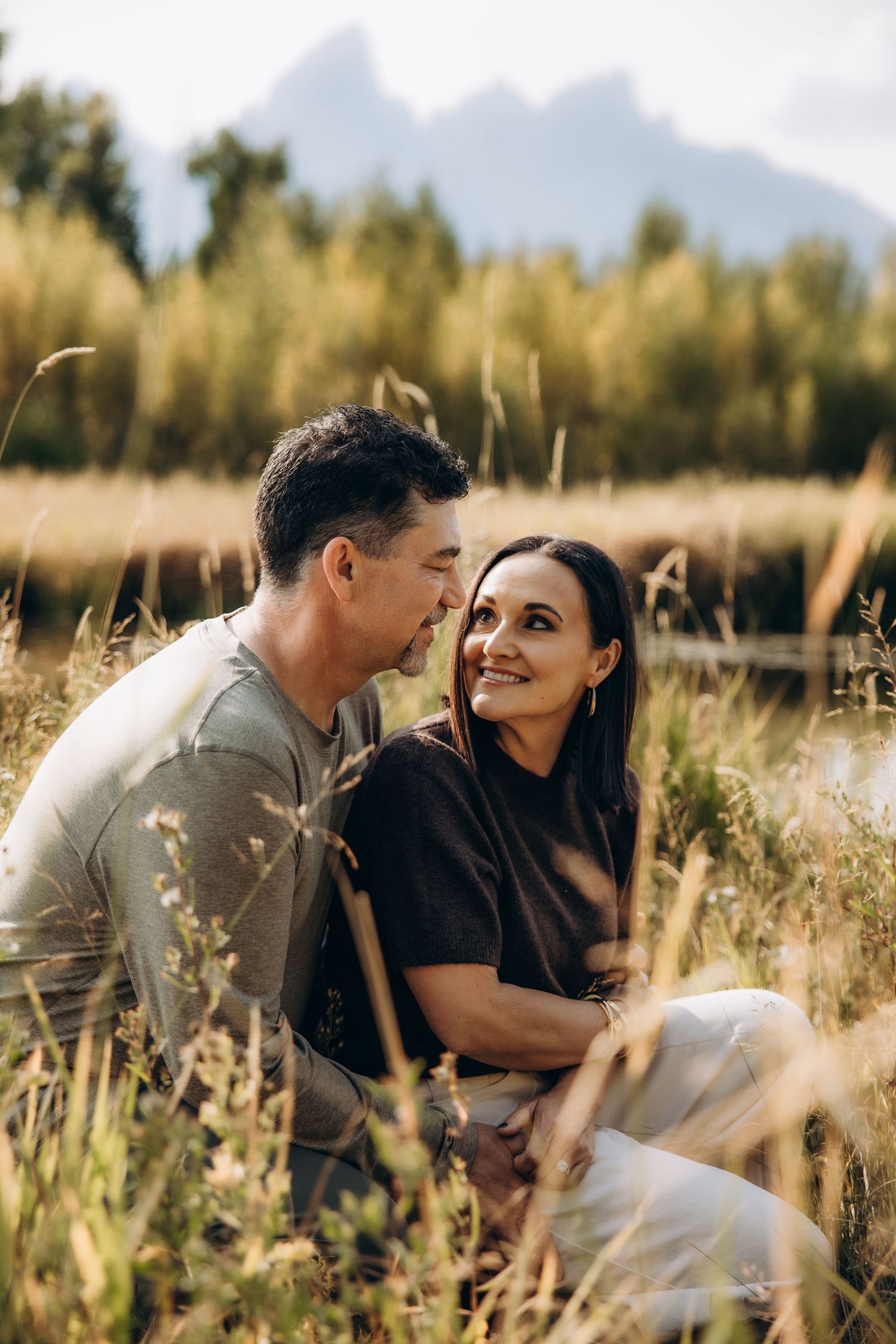 A romantic couple sits together in a golden field of tall grass during sunset while sharing intimate moments.