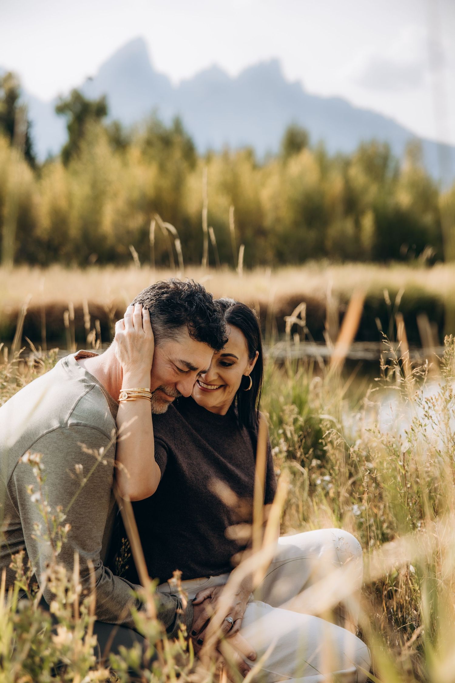 Couple sharing a tender moment lying in tall golden grass with mountains in the background.