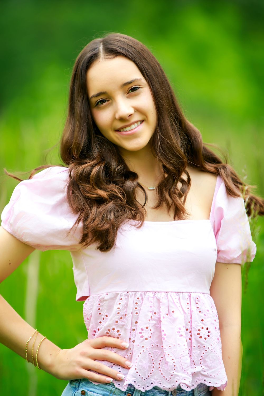 Young woman in pink eyelet top poses against blurred green background in summer photoshoot.