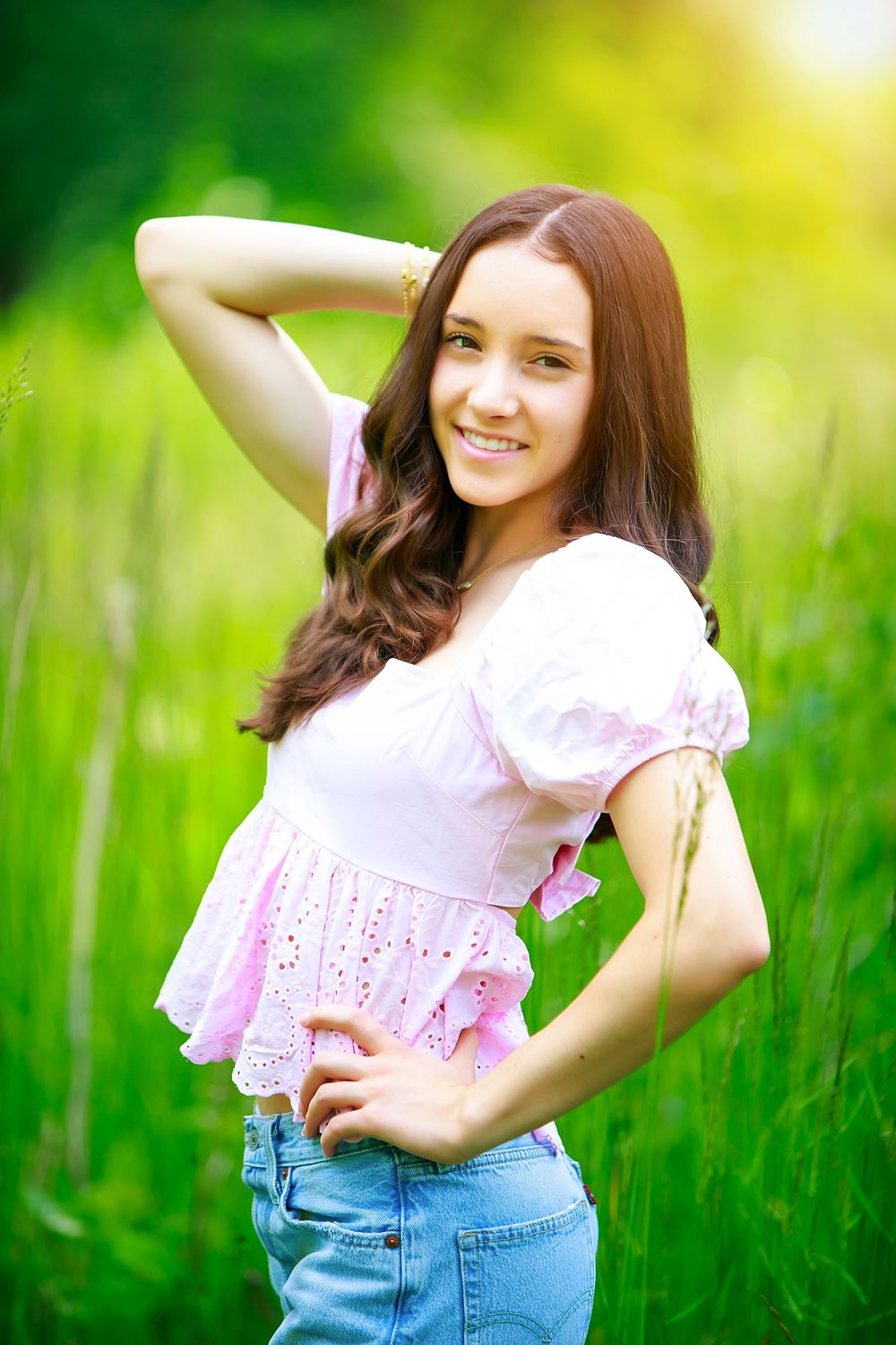 Person in a pink ruffle blouse and jeans poses playfully in a sunlit grass field.