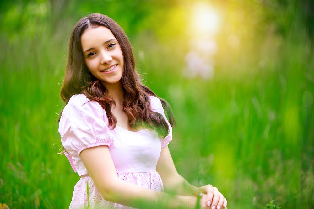 Young woman in pink and white dress posing in sunny outdoor garden setting with vibrant green background.