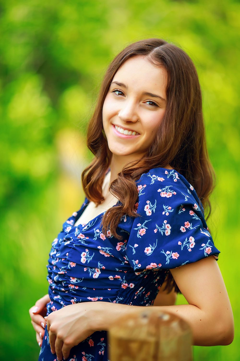 Woman in blue floral dress poses outdoors against blurred green nature background.