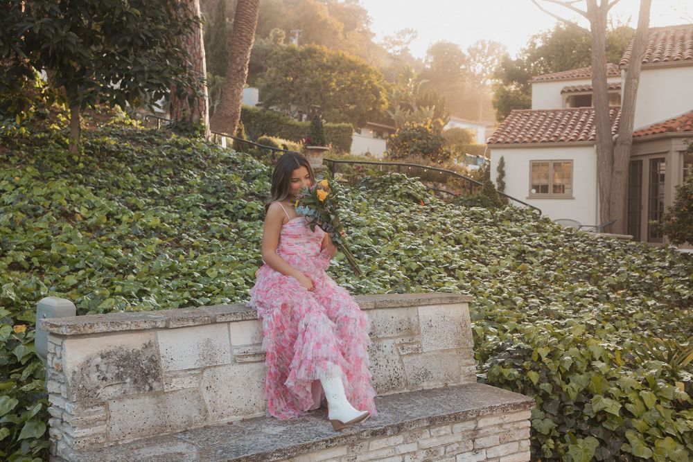 Five serene sunset photos show someone in a pink dress sitting on stone walls amid lush vegetation during golden hour.