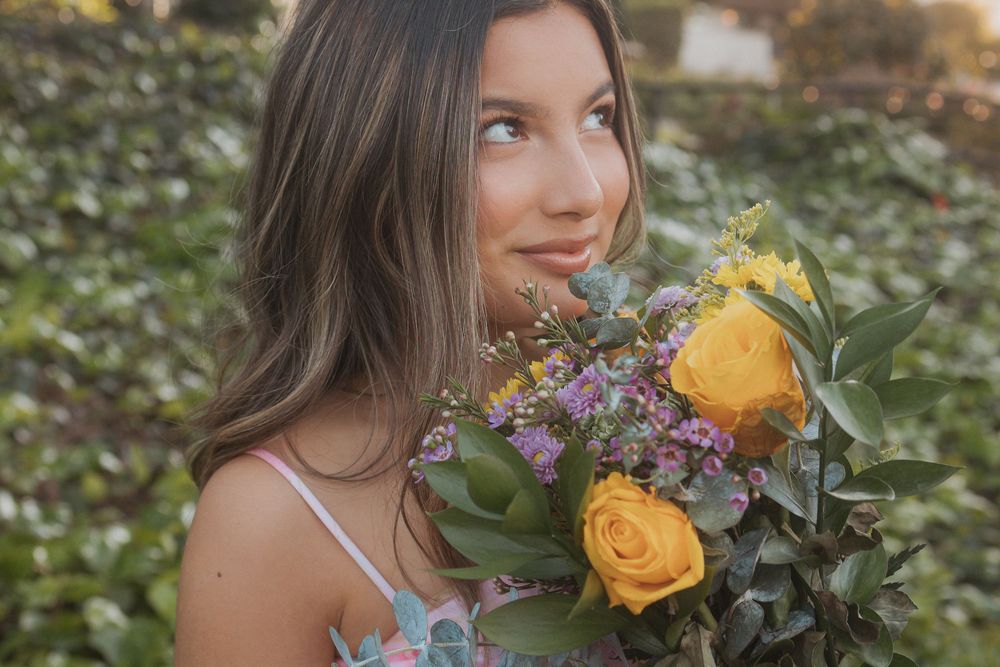 Young woman in light summer dress sits in meadow with yellow roses and wildflowers during warm sunset.