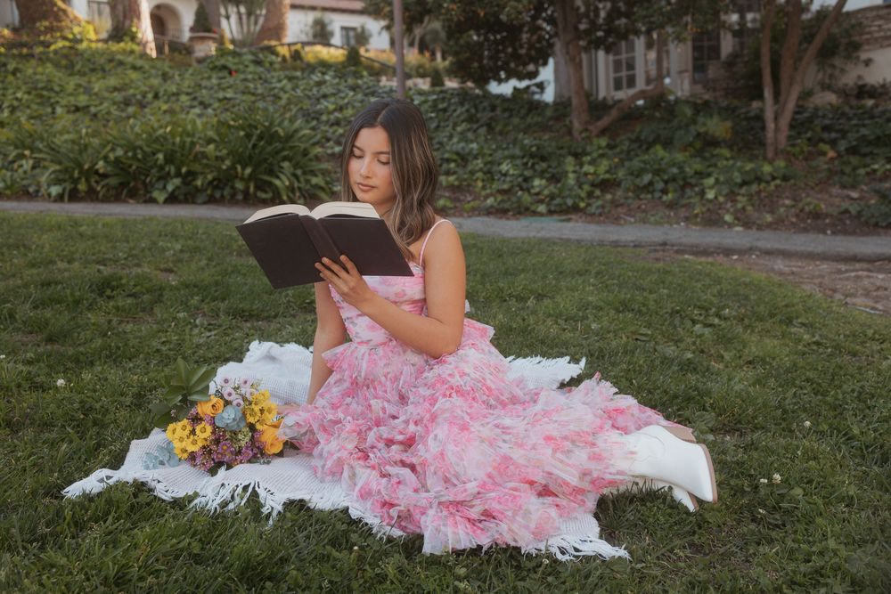 A person in a pink tulle gown reads a book while sitting on a white blanket with flowers in a garden setting.