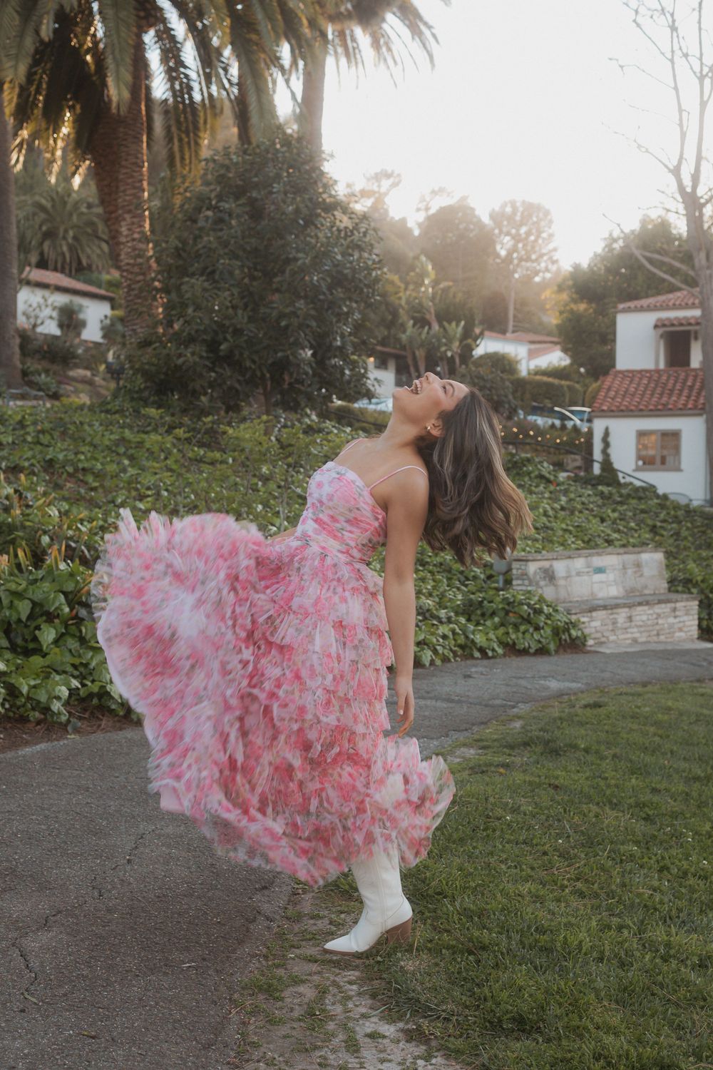 Pink tulle dress with white boots twirls gracefully in a garden setting with houses and trees in the background.