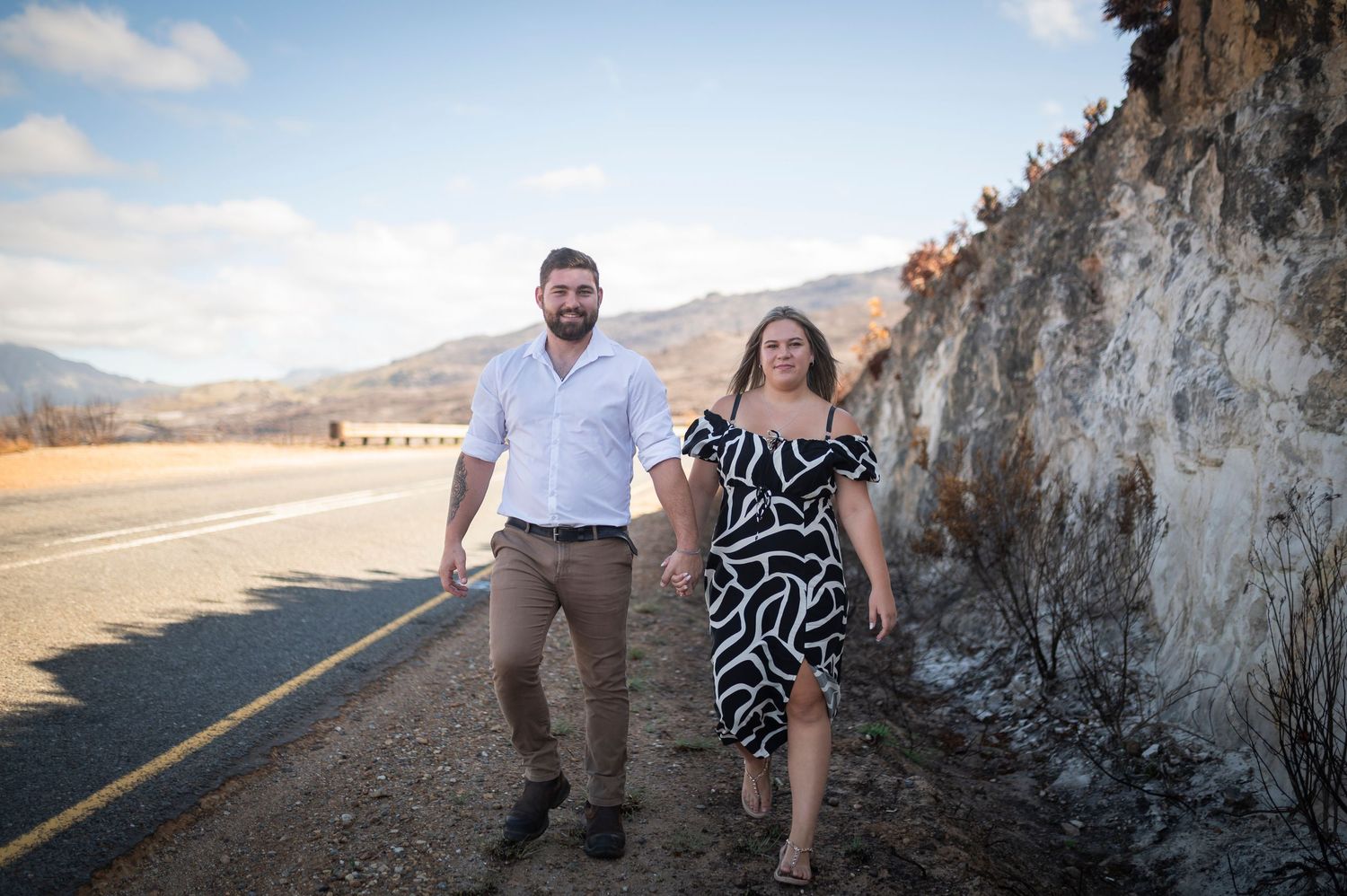 Two people stand against a rocky cliff wall with mountains and desert road in the background at sunset.