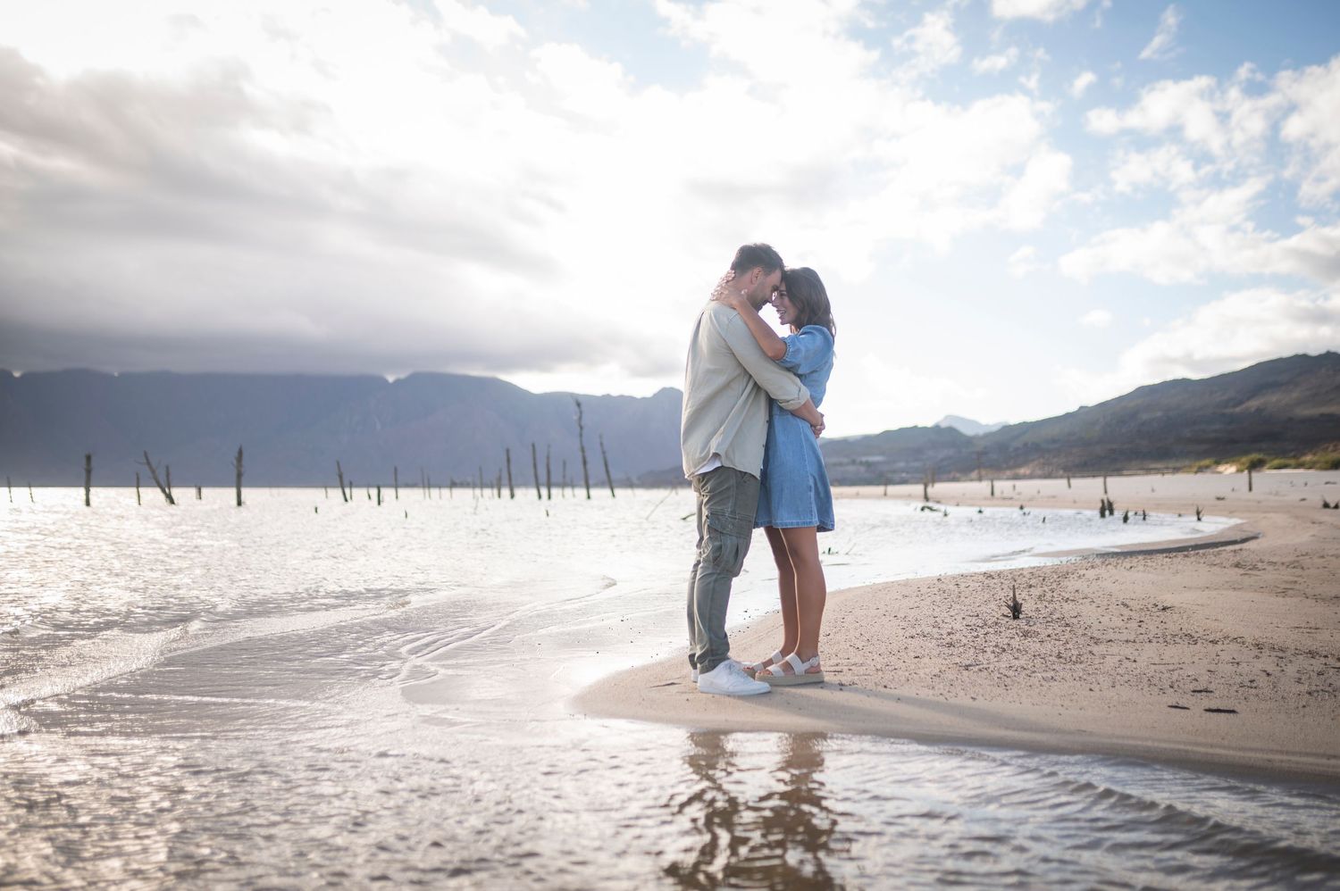 Couple walking together and embracing at sunset on a serene beach with mountains in the background.