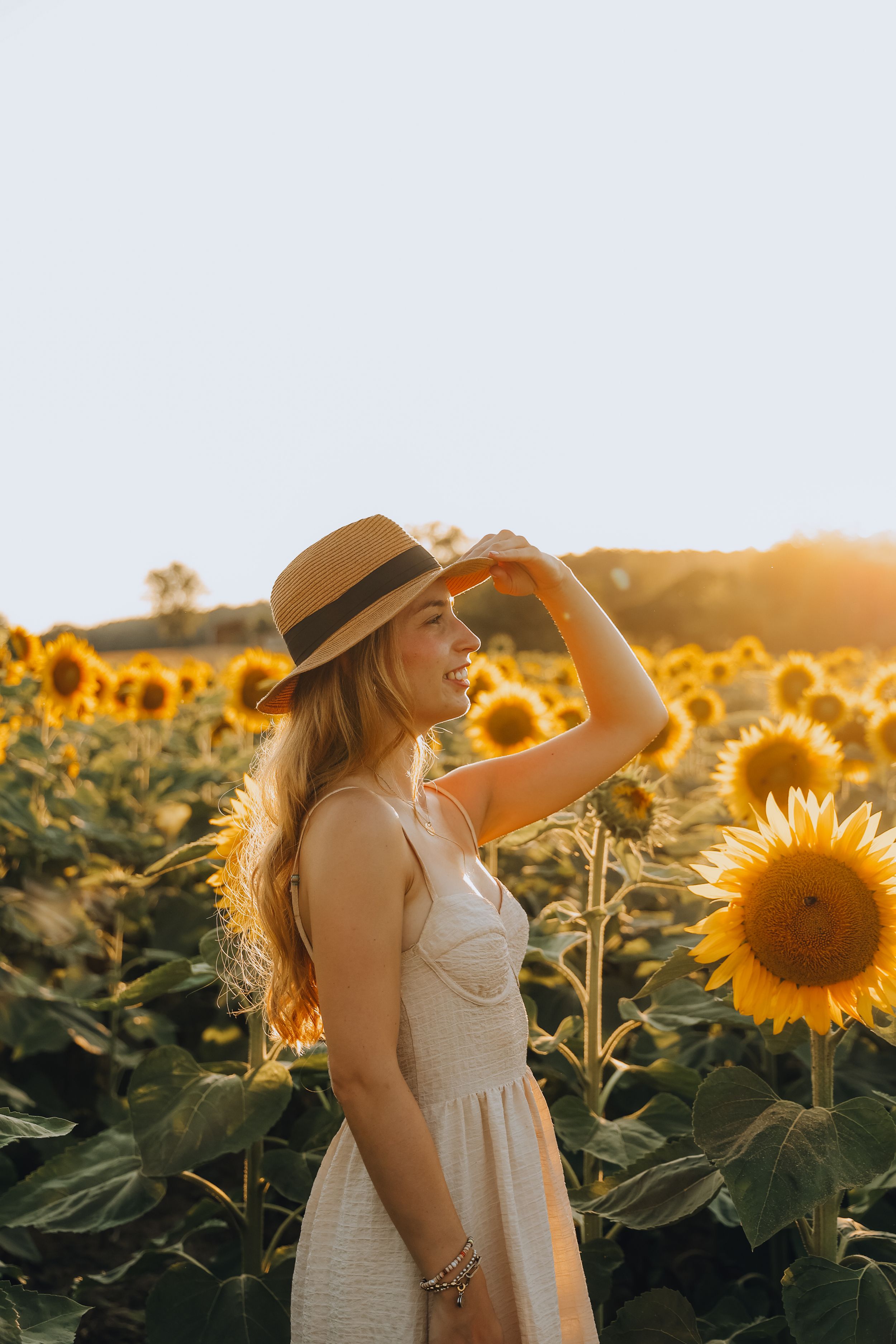 Young woman in white dress and straw hat smiling in a sunflower field at sunset.
