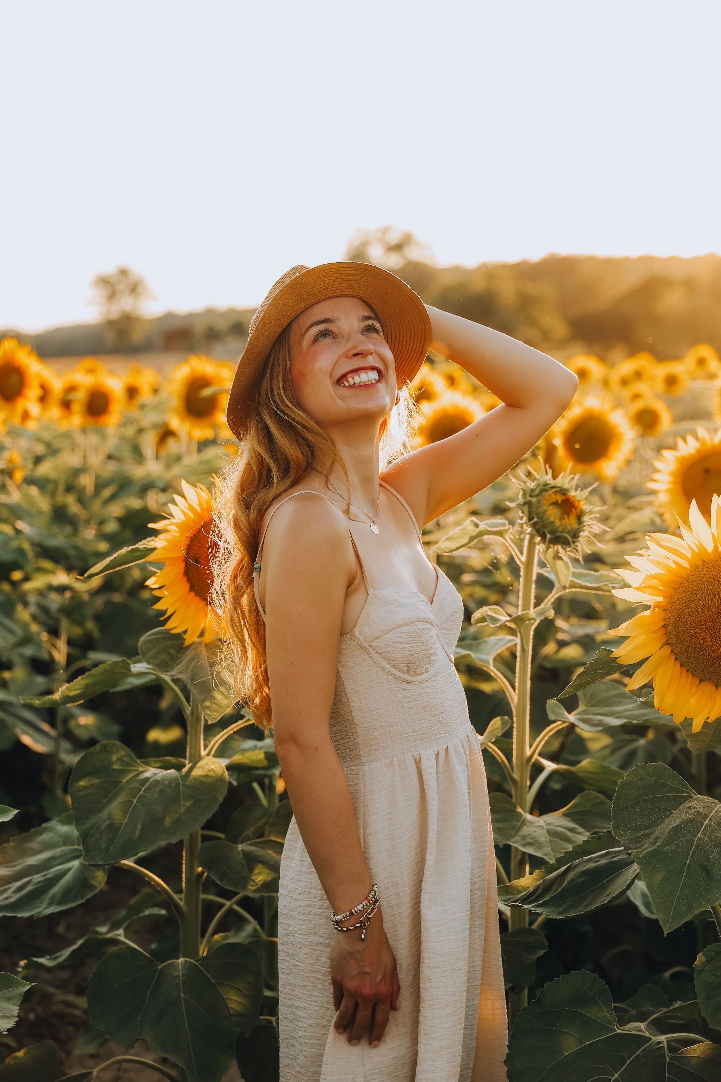 Happy woman in straw hat and white dress laughing in a golden sunflower field.