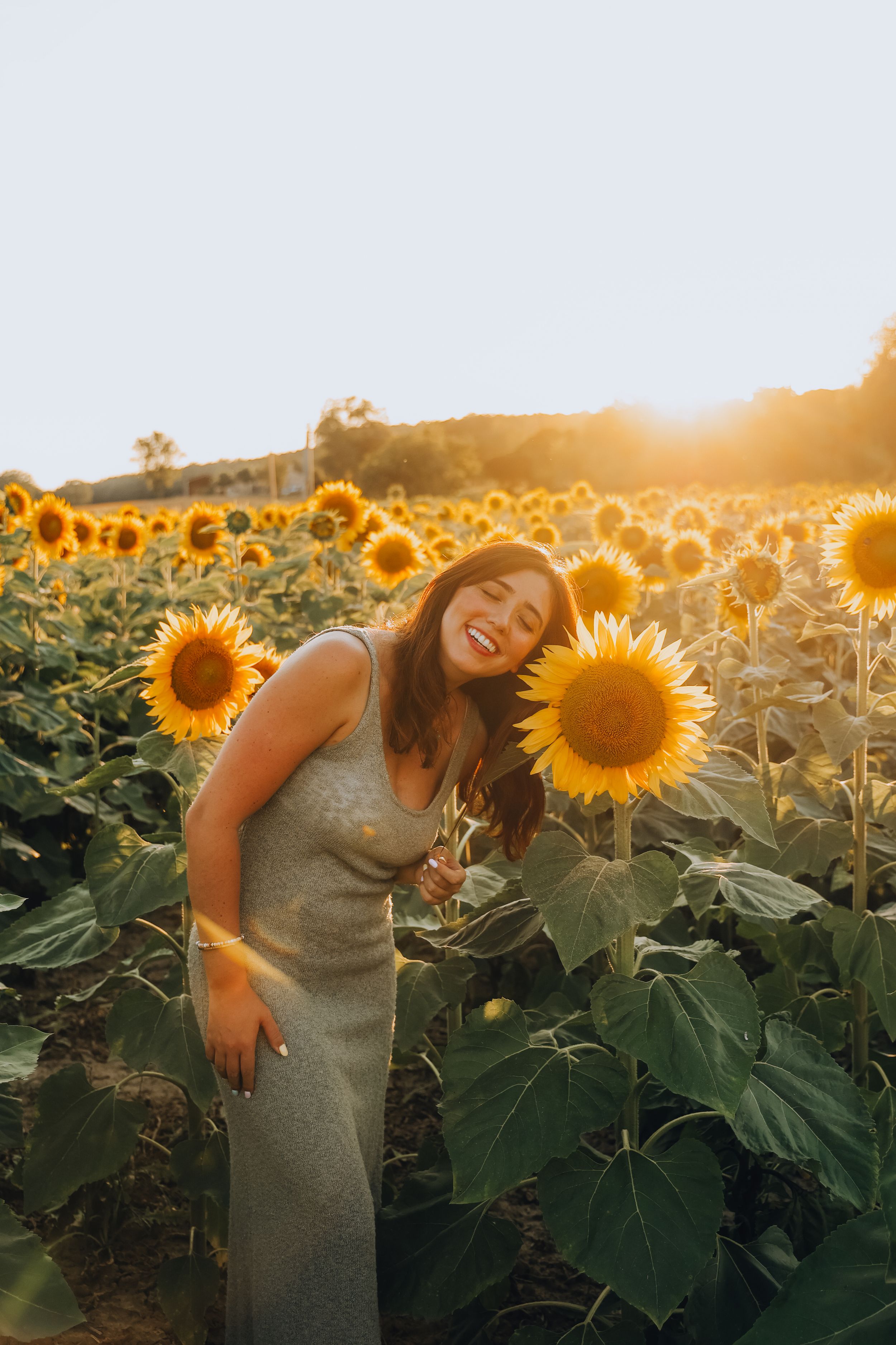 Smiling woman in gray dress leaning toward a sunflower in a glowing sunset field.