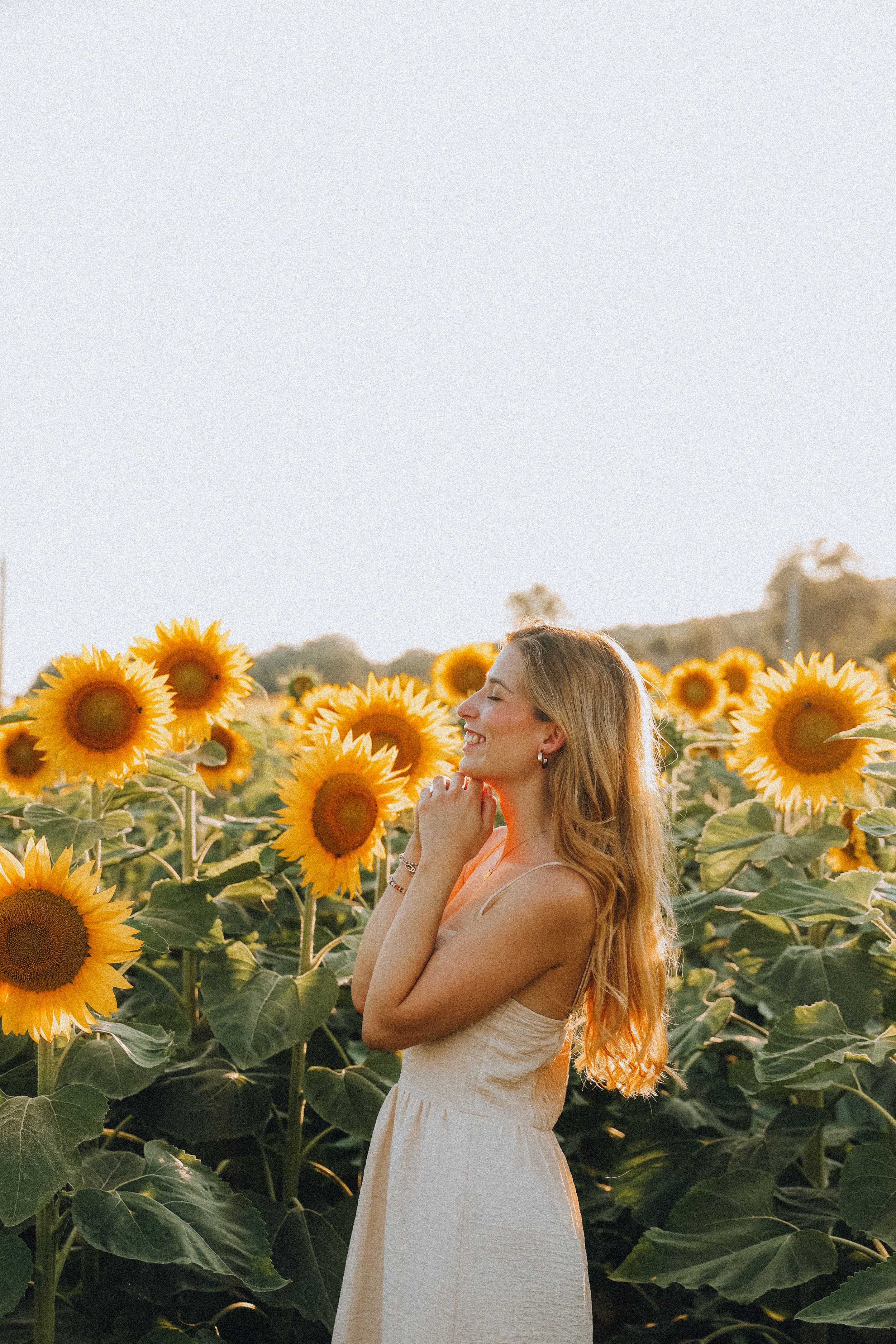 Smiling blonde woman in white dress standing in a sunflower field at golden hour.