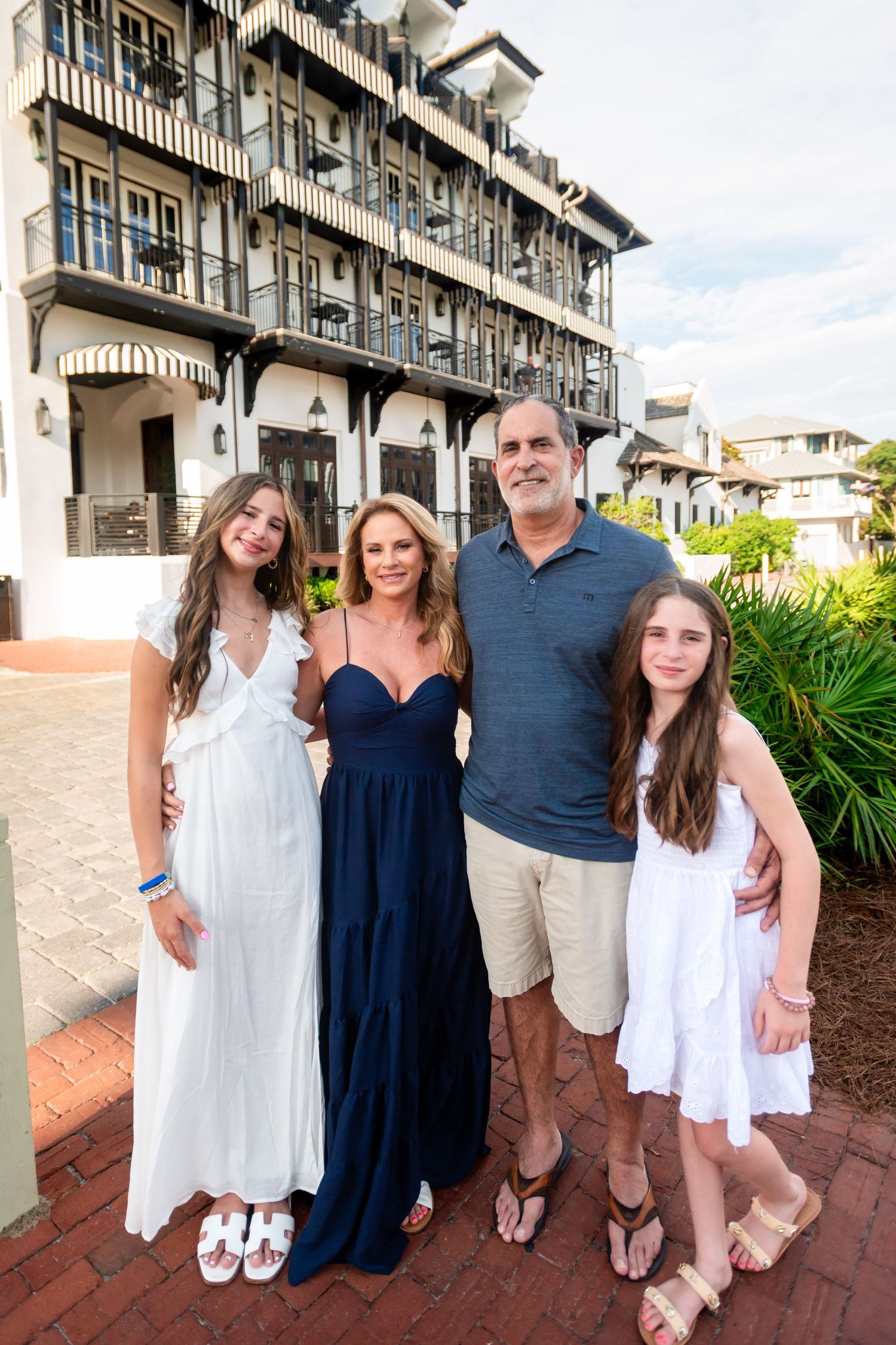 A family poses together on a beachfront balcony wearing coordinated white and navy blue outfits in summer weather.