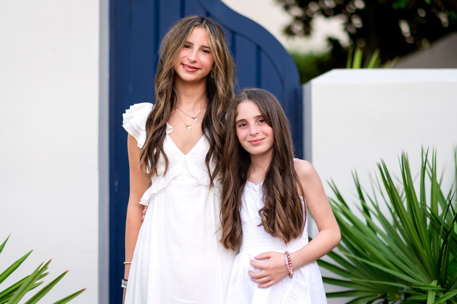 Two people pose together outdoors by a palm plant near a white building exterior.