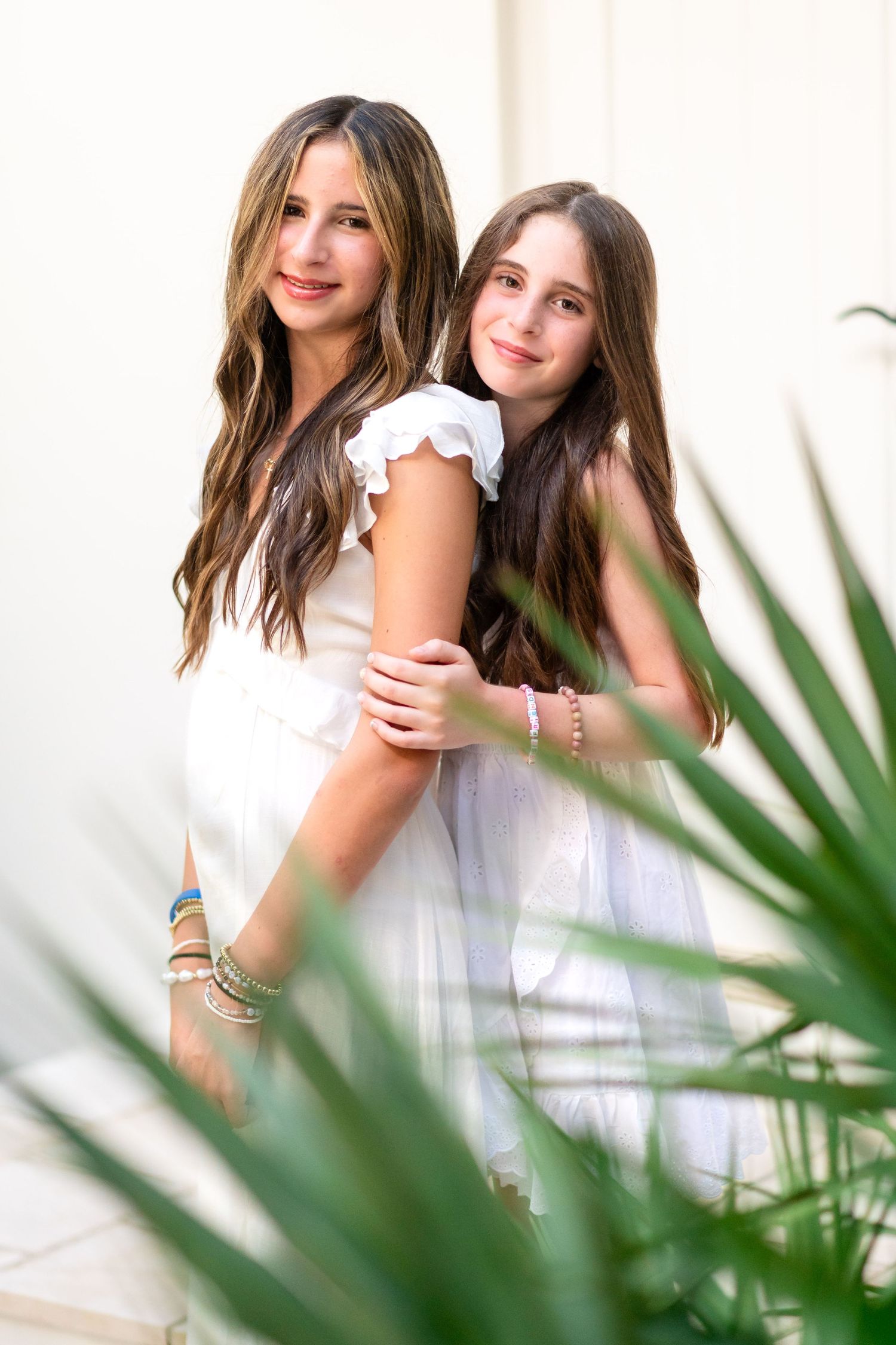 Two people in white summer dresses standing close together while smiling in front of tropical foliage.