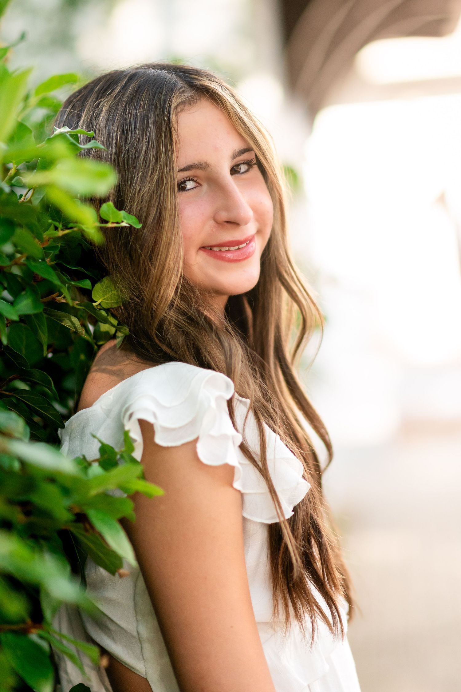 Woman in white ruffled dress poses against blurred tropical background with charming smile.