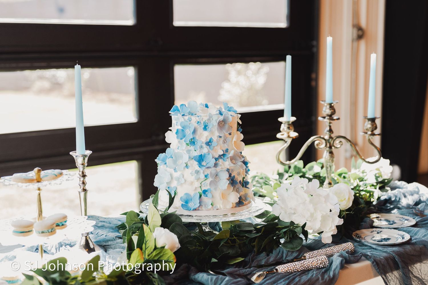 A white wedding cake decorated with blue confetti-style accents sits on an elegant table with candles and floral arrangements.