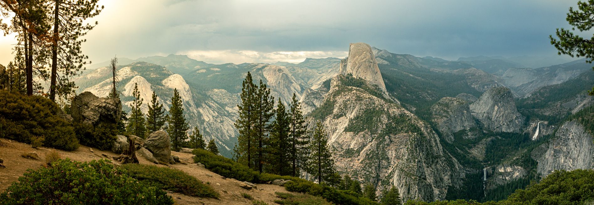 Panoramic view of Yosemite Valley with Half Dome, pine trees, and dramatic stormy skies at sunset.