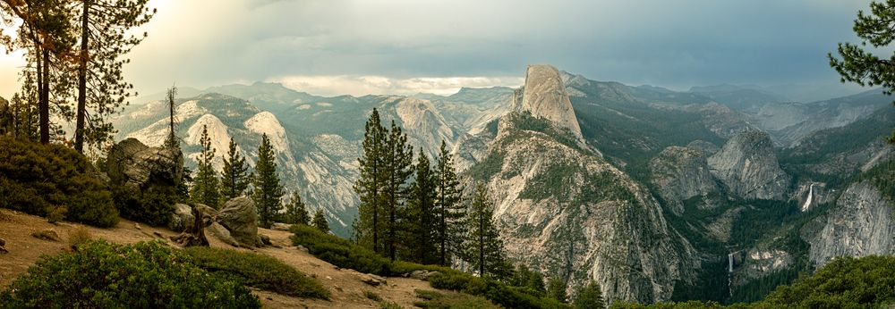 Panoramic view of Yosemite Valley with Half Dome, pine trees, and dramatic stormy skies at sunset.