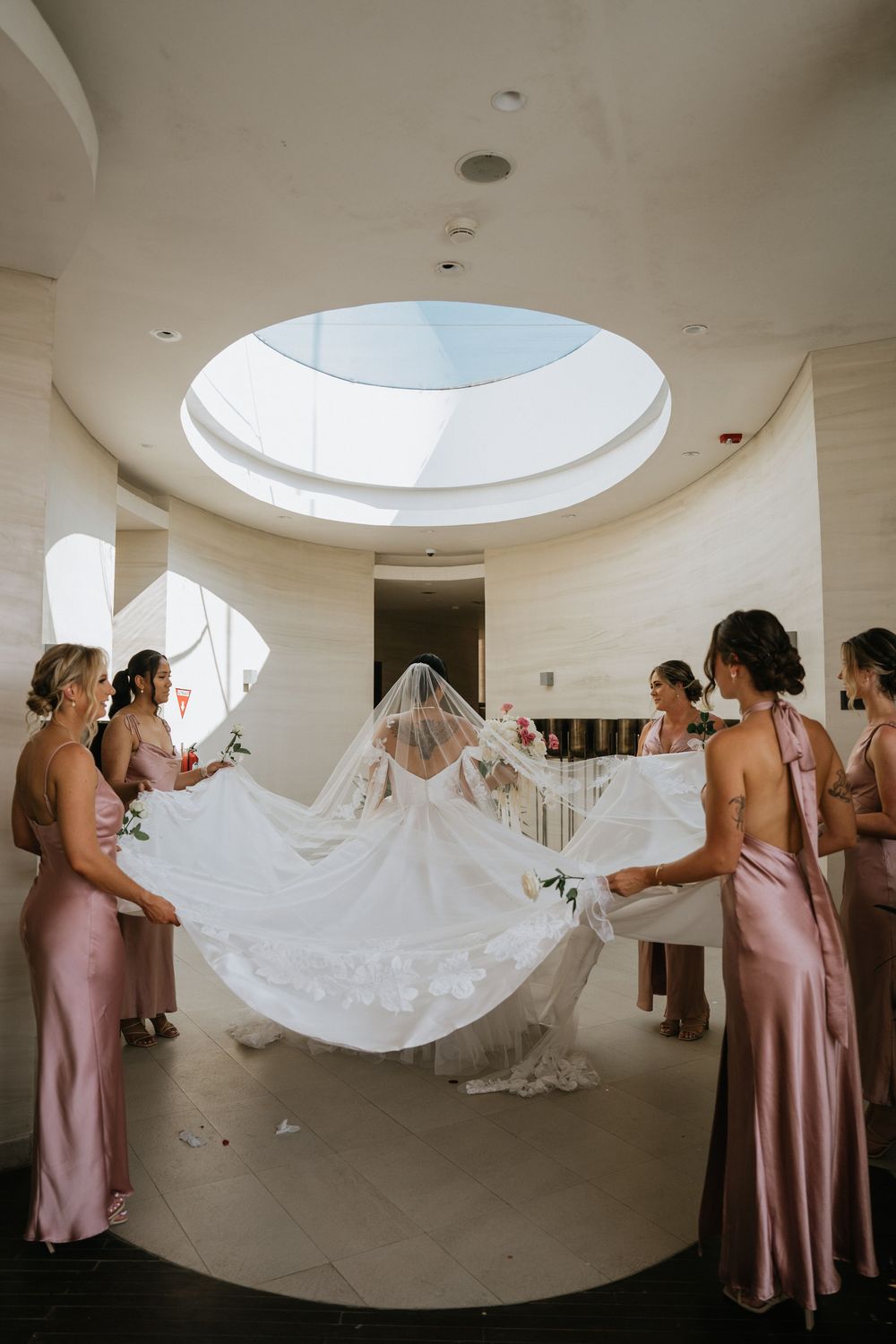 Bridesmaids in pink dresses help arrange bride's veil under circular skylight.