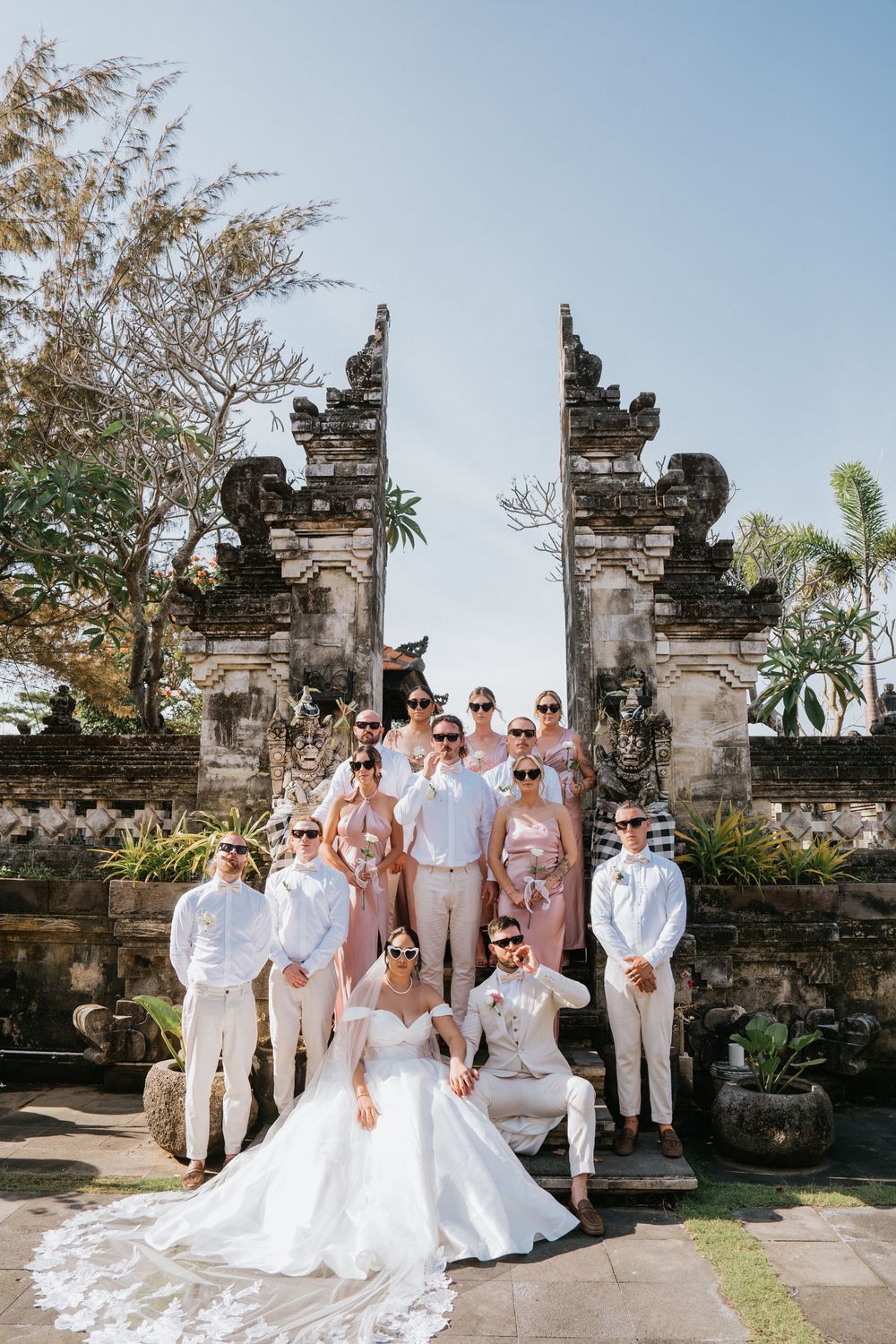 Wedding party poses in front of traditional Balinese stone temple gates wearing white and beige formal attire.