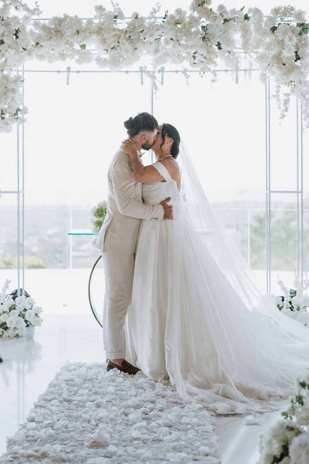 Romantic wedding couple shares intimate moments under floral archway with white petals scattered on floor.