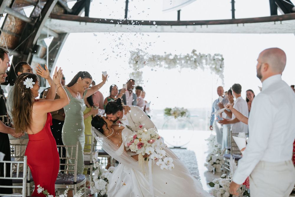 Wedding reception guests raise glasses in celebration during indoor party with string lights.