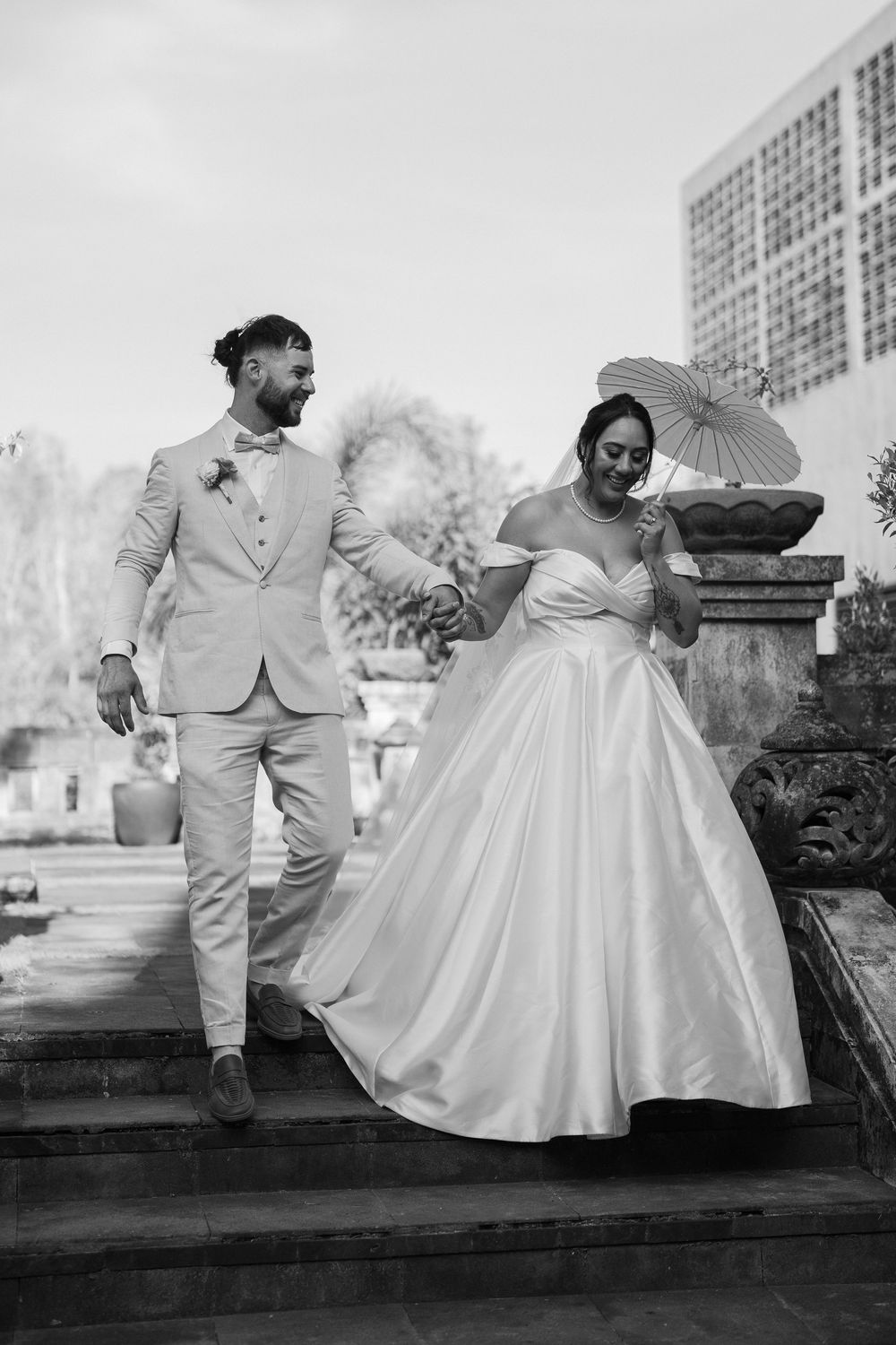 Black and white portrait of couple in wedding attire posing on stone steps with parasol.