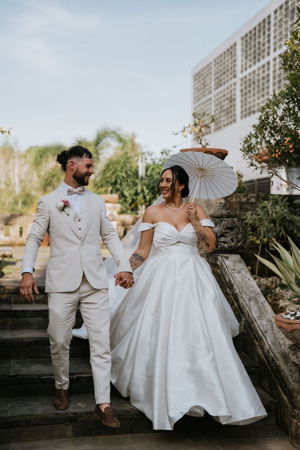 Couple in wedding attire walks along stone wall pathway holding hands and parasol.